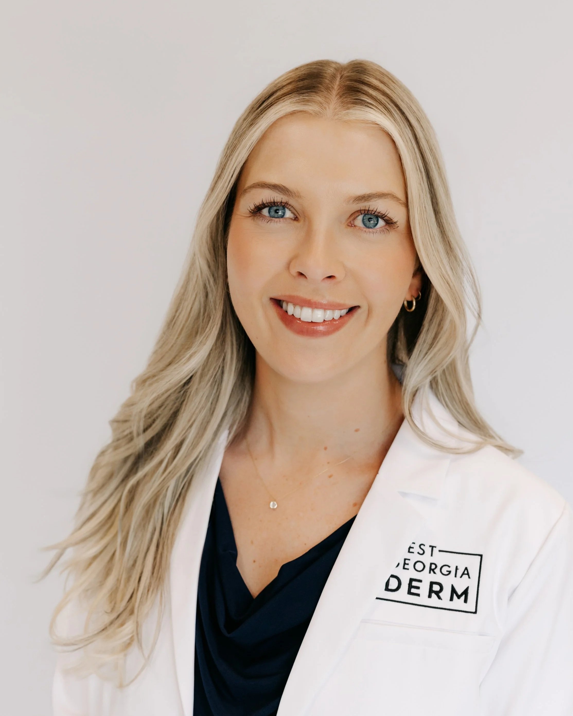 A smiling woman in a white lab coat with a badge that reads 'West Georgia Derm.'