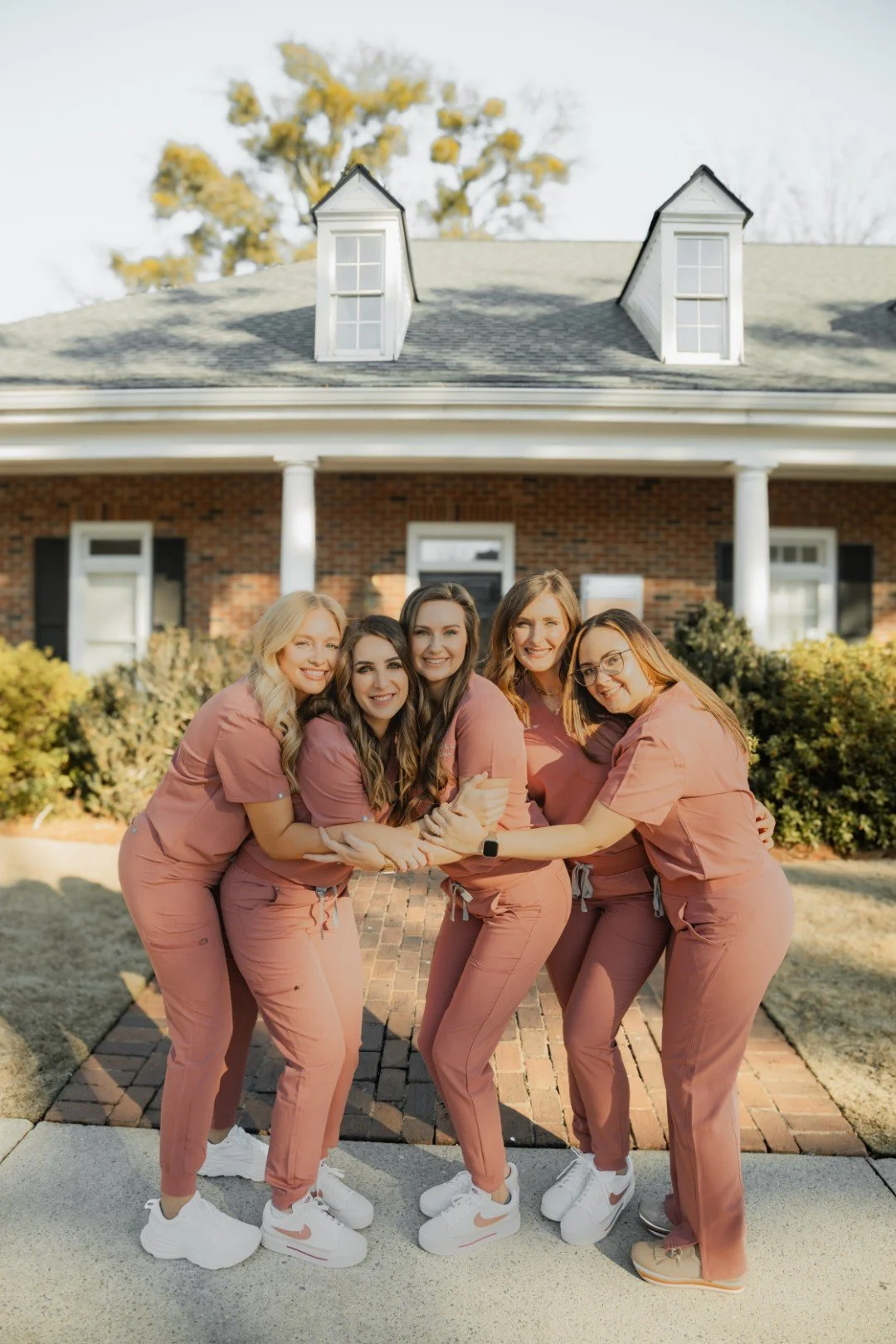 Five women in matching pink scrubs and white sneakers smiling and embracing in front of a brick building with a porch and dormer windows.
