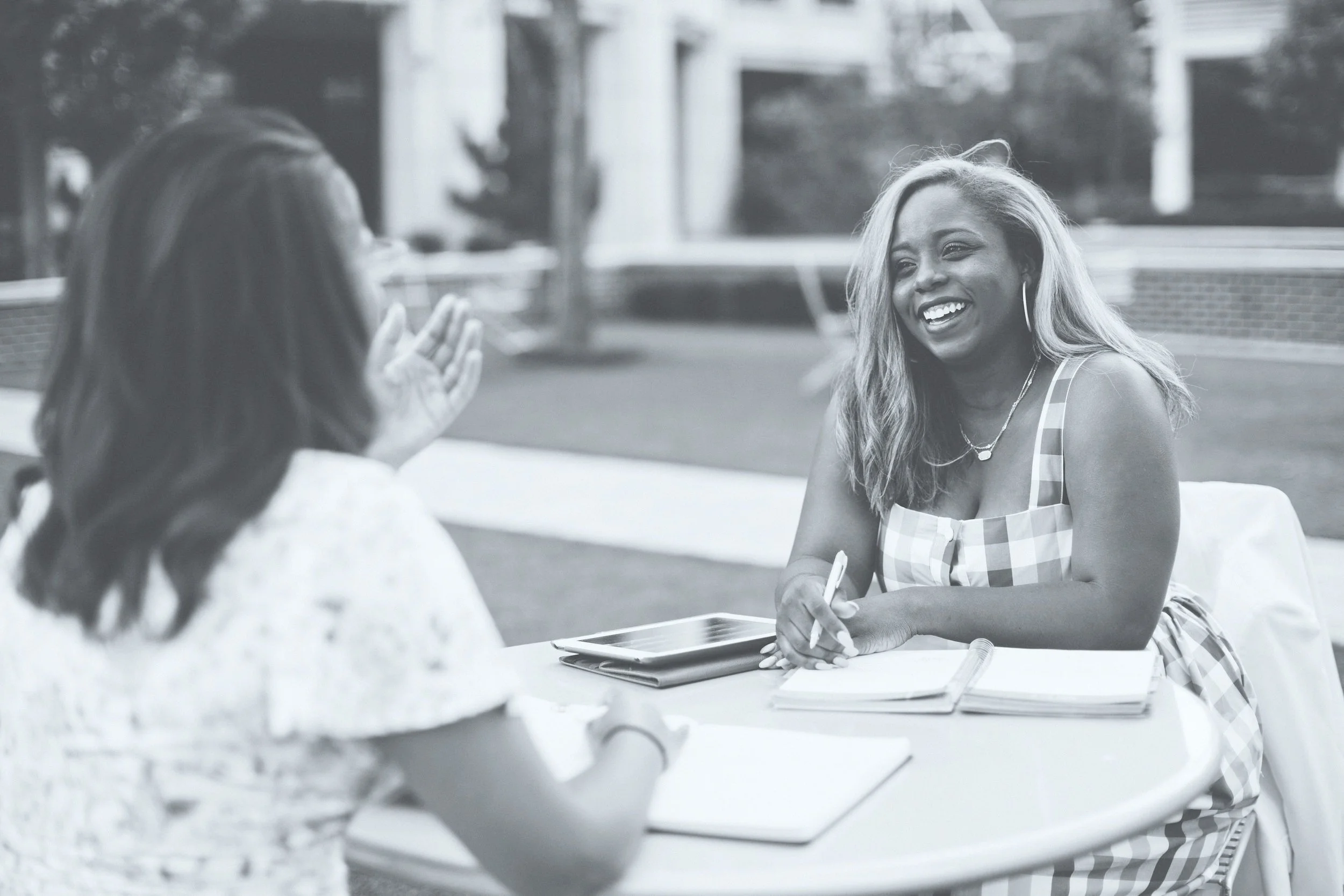 Two women engaged in conversation at an outdoor table with notebooks and a tablet, smiling and enjoying each other's company.