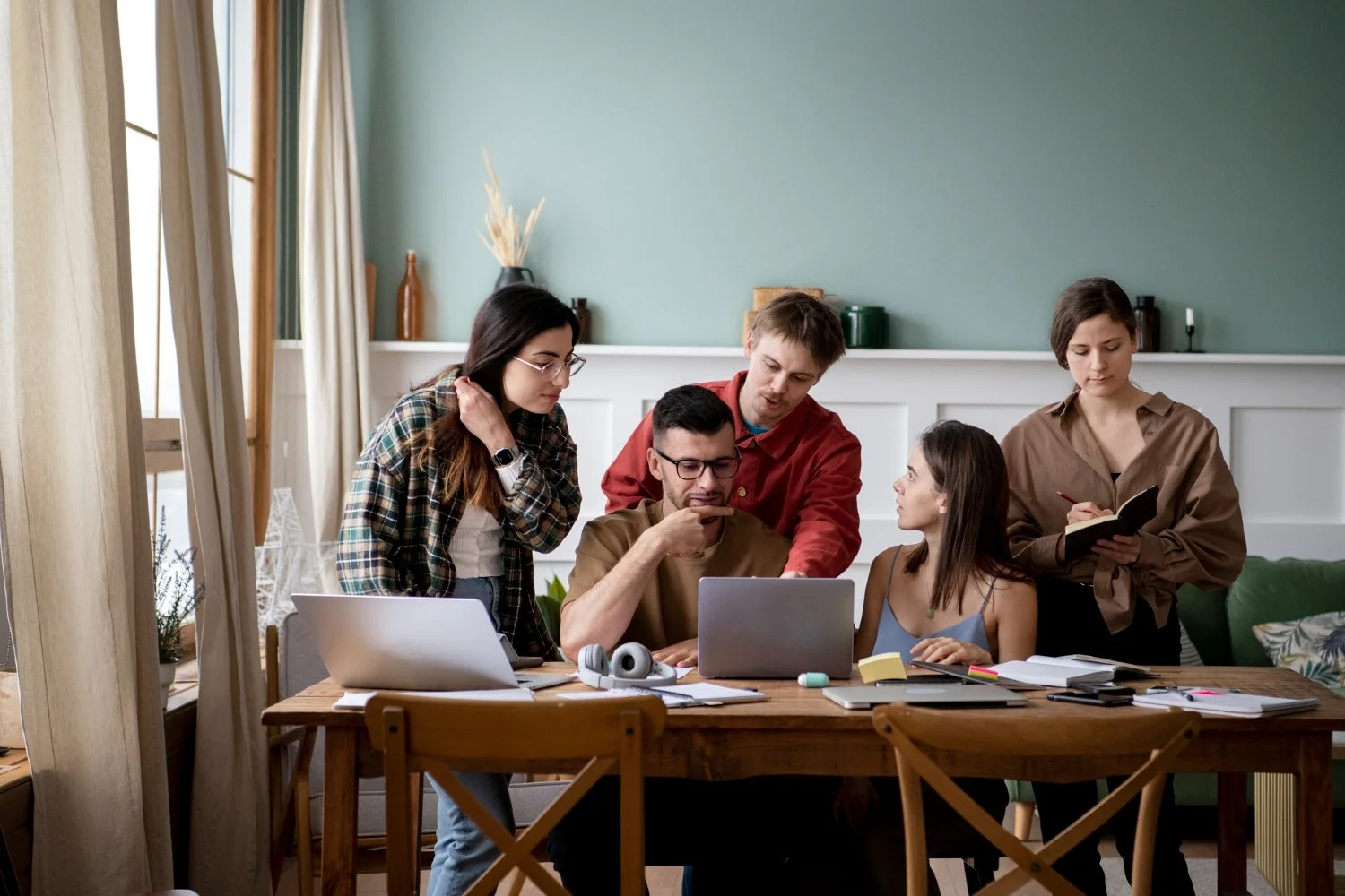 Six people gather around a wooden table working on a laptop, with papers and notebooks, in a room with green walls and curtains.