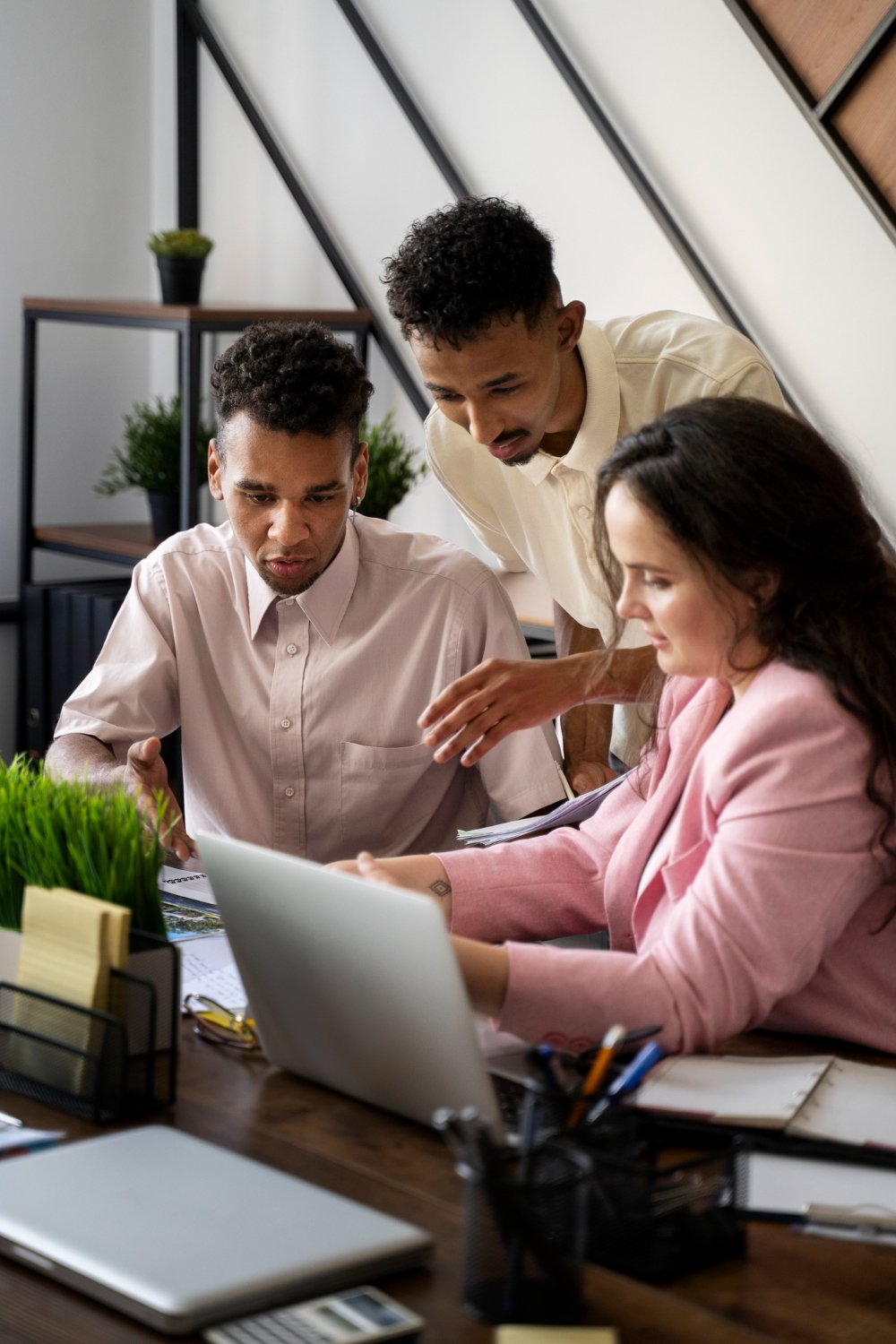 Three colleagues working together at a desk in an office, reviewing documents on a laptop.