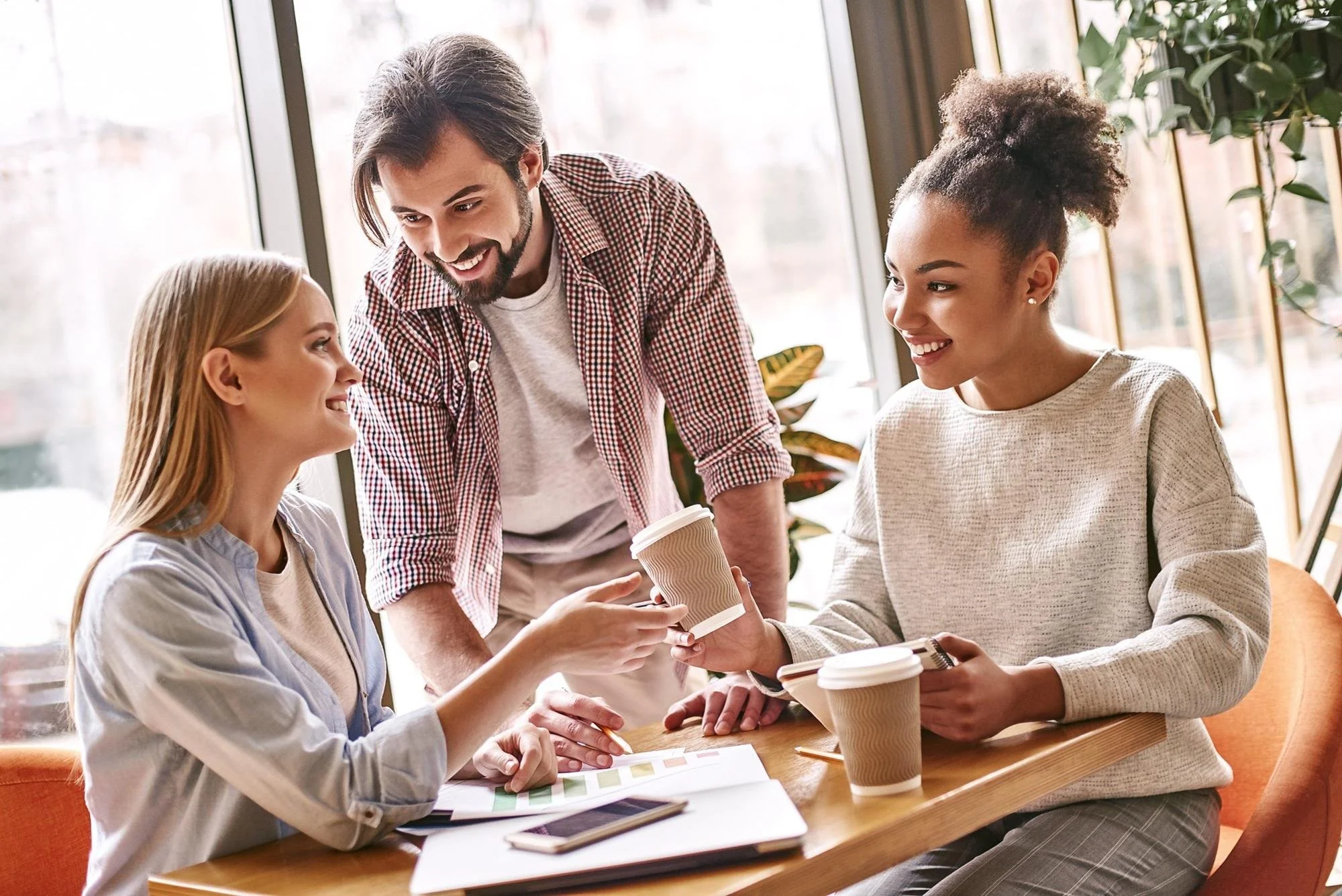 Three women and a man engaged in a discussion over coffee in a bright cafe with large windows and plants.
