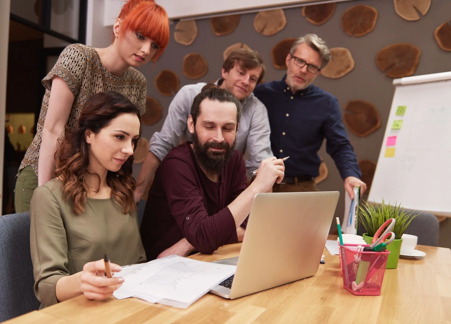Five colleagues gathered around a table, looking at a laptop and discussing a project in an office setting, with a whiteboard and decorative wall behind them.