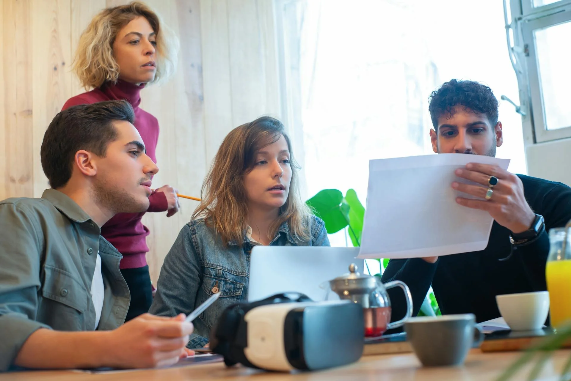 Group of four young adults gathered around a table, with one man holding a sheet of paper and another person pointing at it, in a room with wooden walls and natural light.