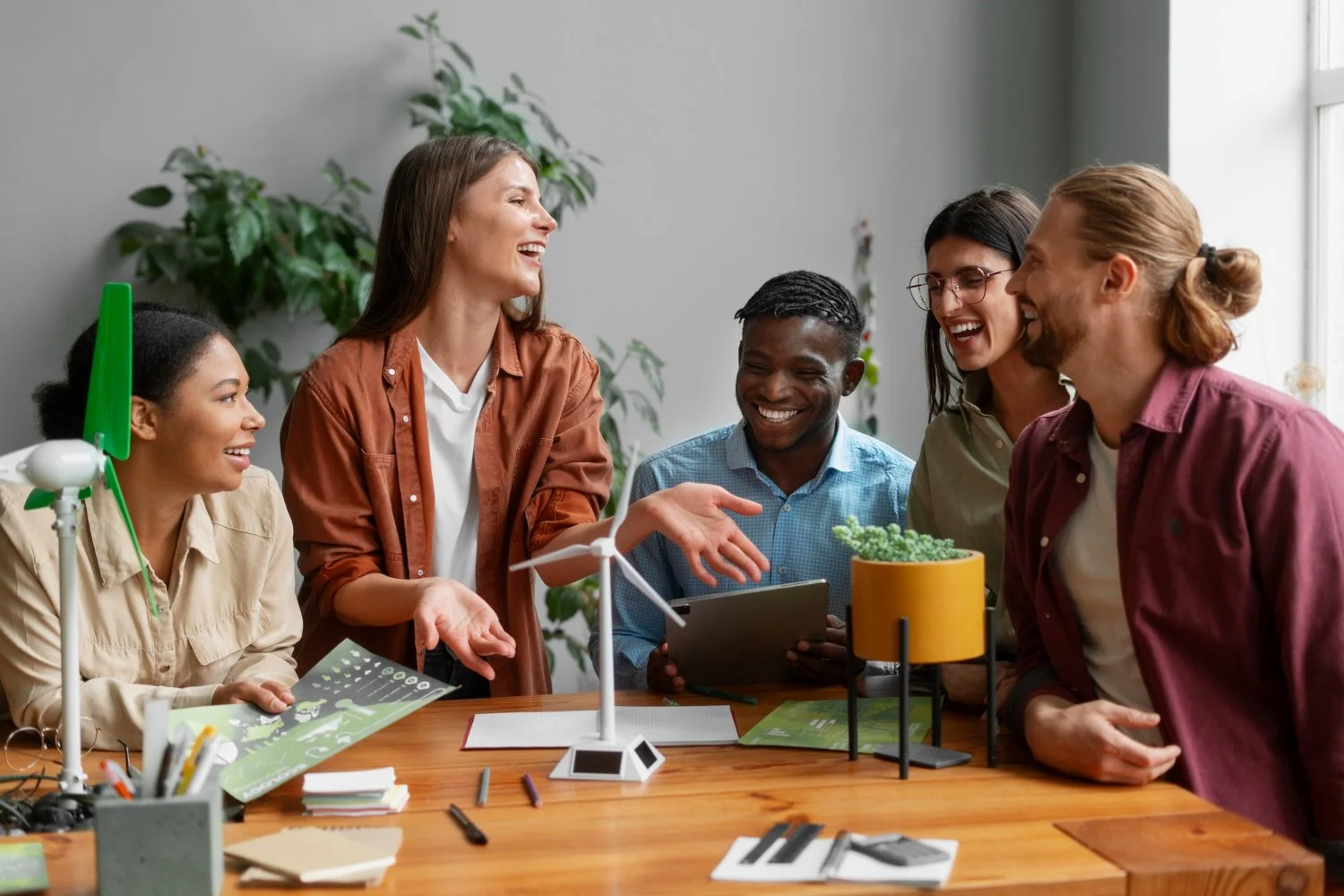 Group of five young adults smiling and talking around a wooden table in an office, with a model wind turbine and green plants, engaging in a lively discussion about renewable energy.