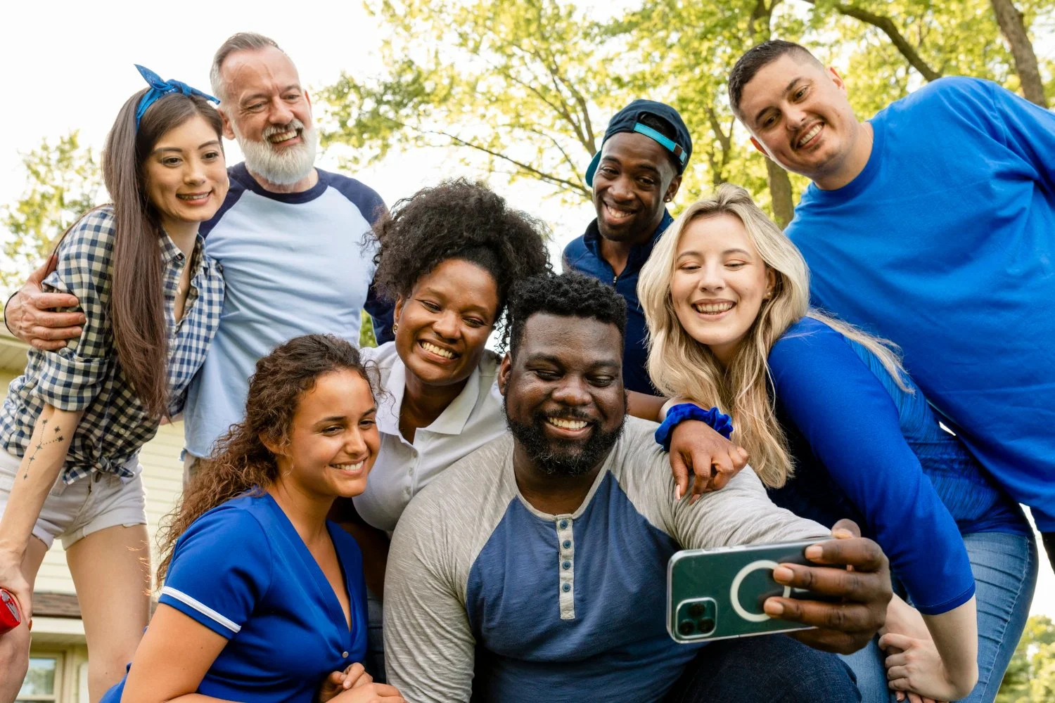 Group of ten smiling people, diverse ages and ethnicities, taking a selfie outdoors with trees and a house in the background.