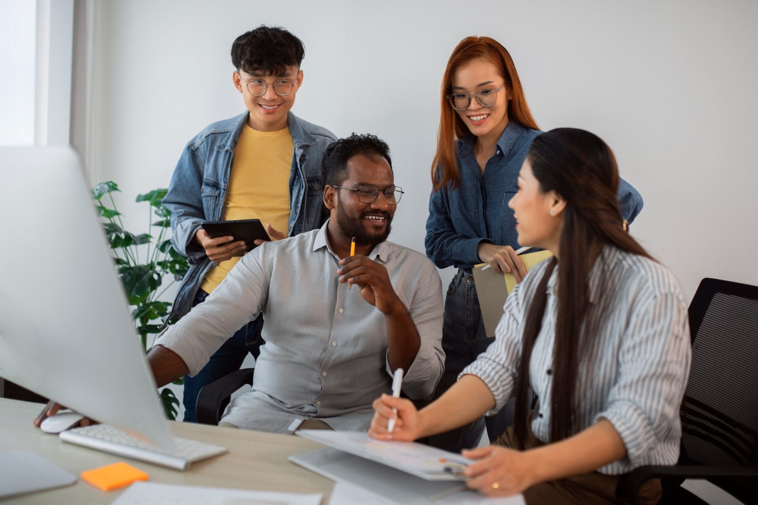 Four diverse coworkers in an office, engaging in a discussion around a computer monitor, with one seated woman writing, two standing women smiling, and a seated man holding a pencil and looking at her.