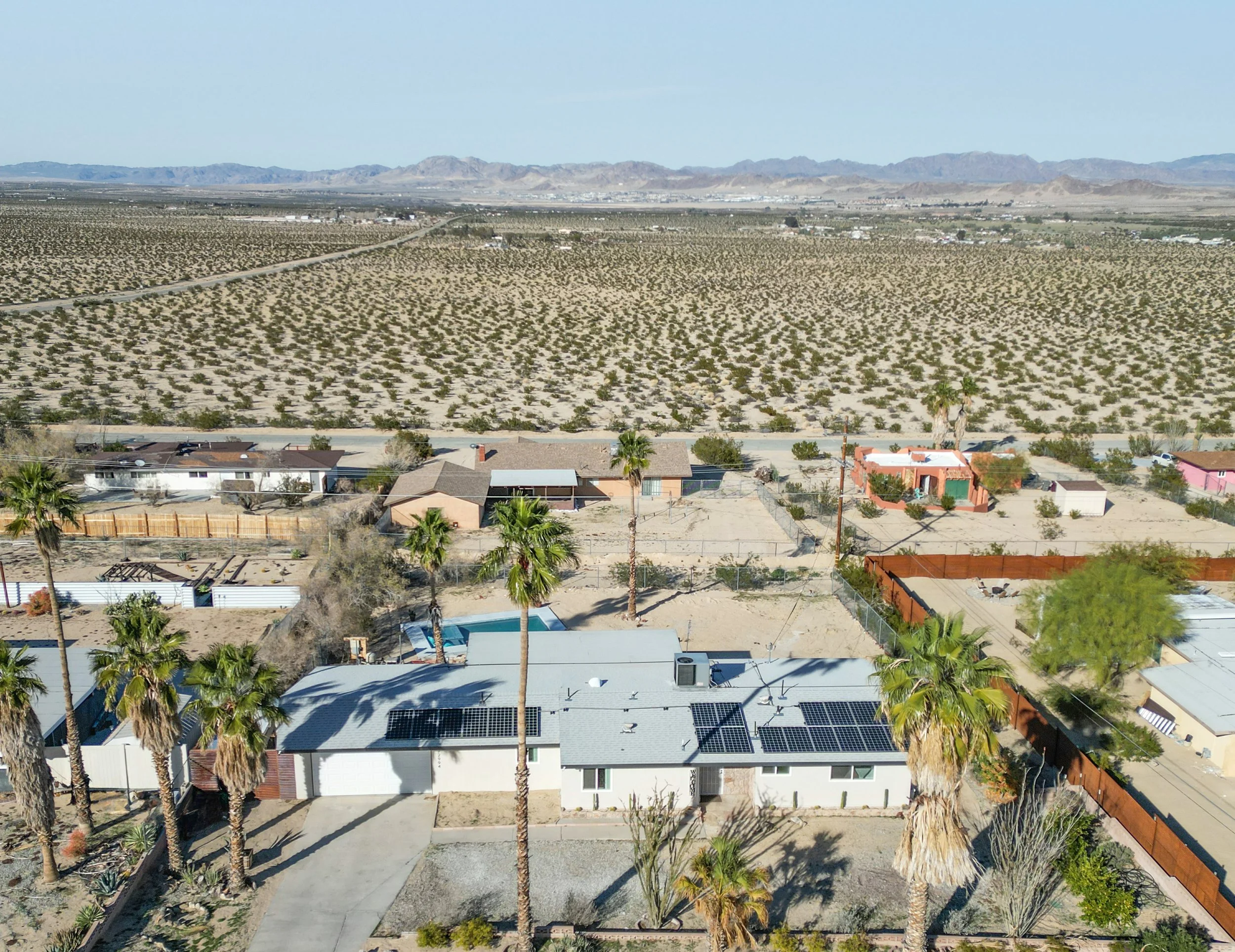 A desert landscape with suburban houses in the foreground, containing solar panels on rooftops, and a vast desert with sparse vegetation and mountains in the background.