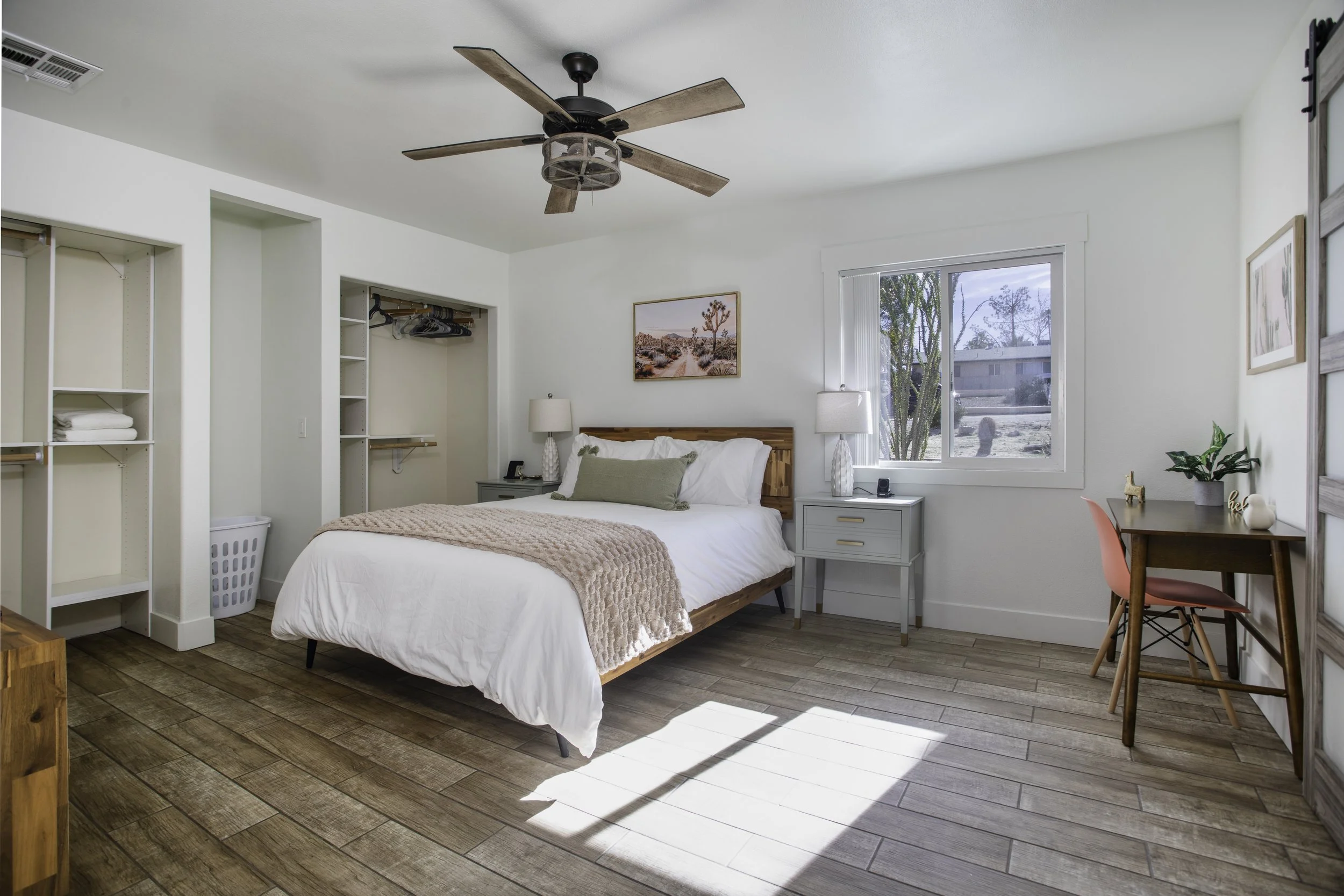 A bedroom with a bed, two nightstands with lamps, and a walk-in closet. There is a window with sunlight, a ceiling fan, and a small desk with a pink chair and plants.