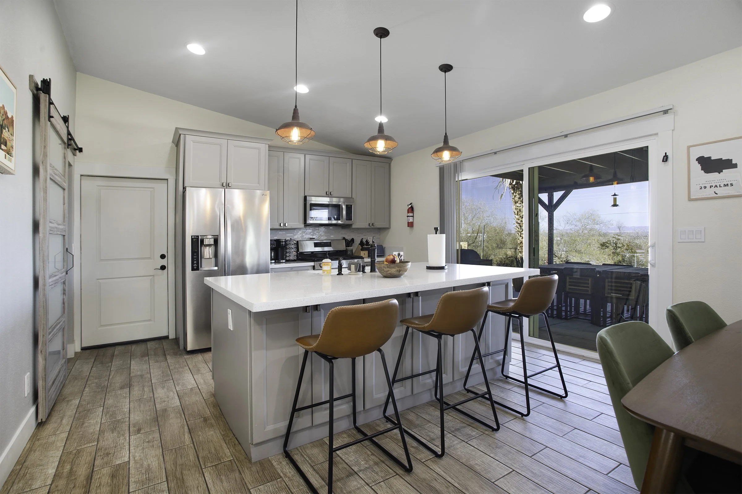 Modern kitchen with gray cabinets, stainless steel refrigerator, microwave and oven, white island with three brown chairs, wooden flooring, sliding glass door leading to an outdoor patio with a view of trees.