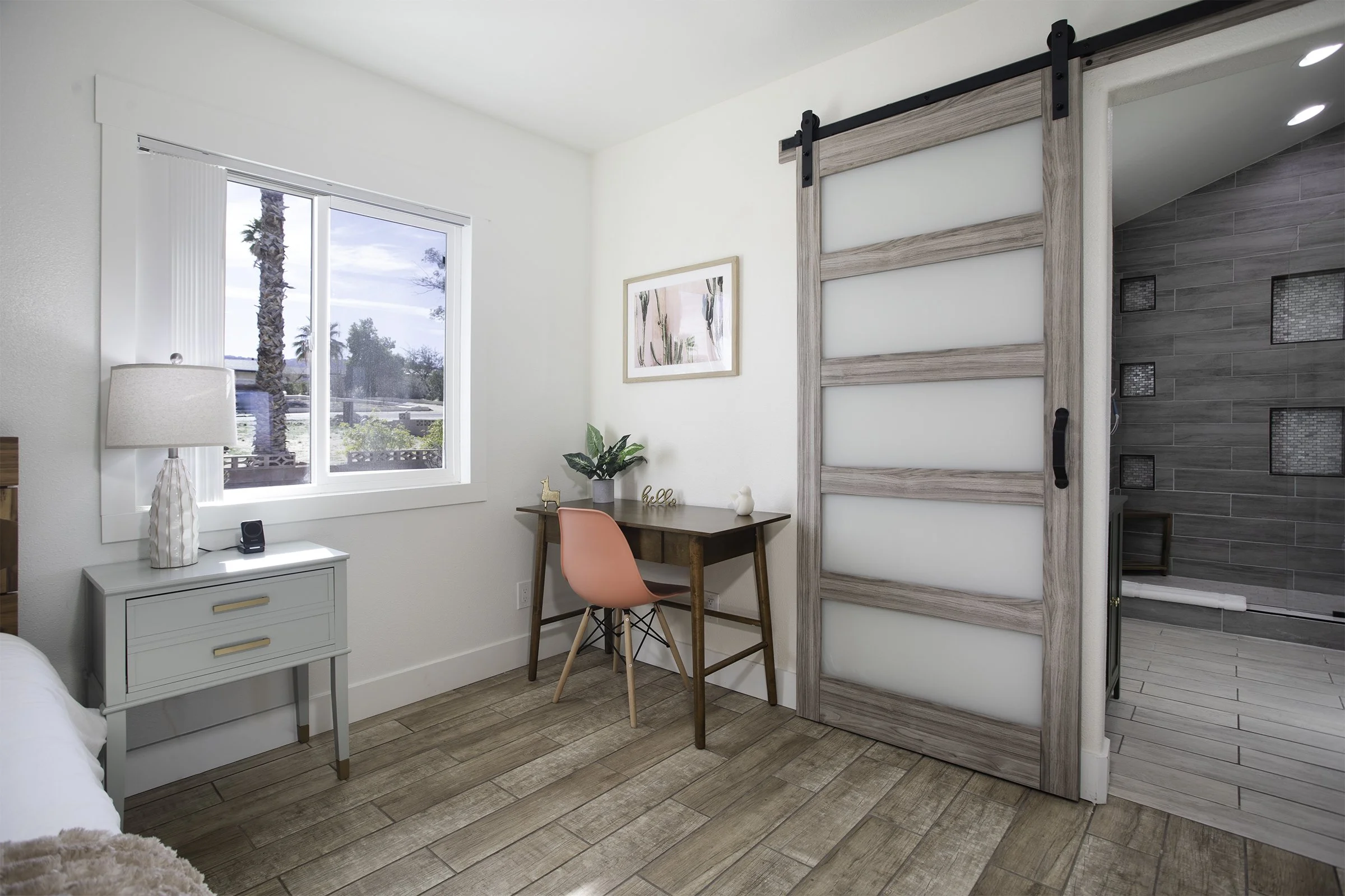 A cozy bedroom corner with a small white nightstand, a large window with vertical blinds, a wooden desk with a pink chair, a potted plant, and a sliding barn door leading to a bathroom with gray tiled walls.