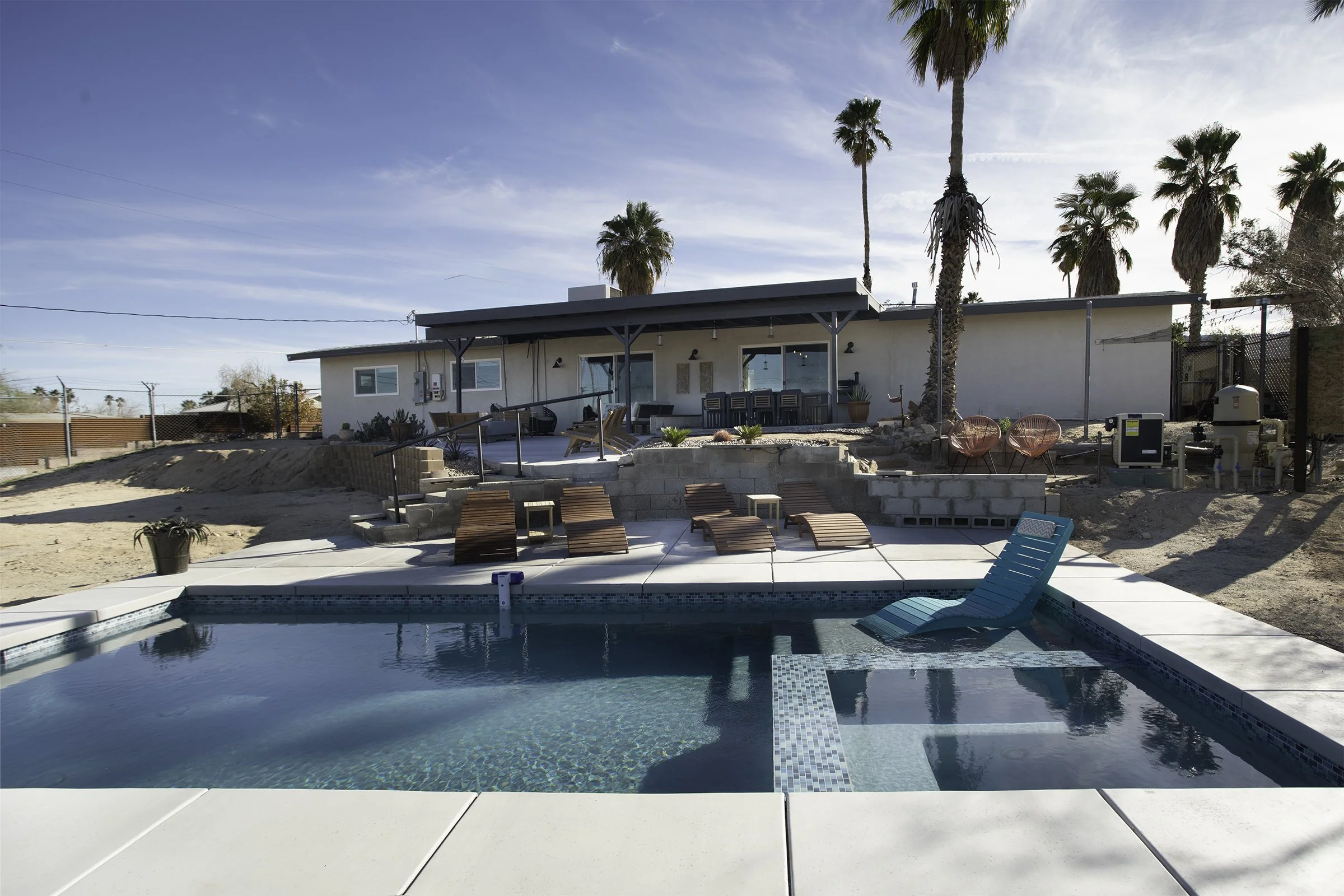 An outdoor backyard with a swimming pool, lounge chairs, potted plants, and a house with a patio and palm trees under a partly cloudy sky.