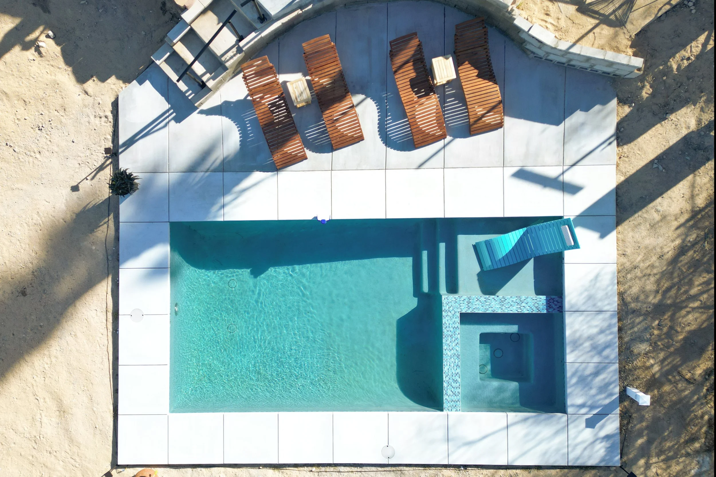 An aerial view of a pool area with a swimming pool, a small hot tub, a blue lounge chair, several wooden lounge chairs, and a tiled patio surrounded by dirt and sand.