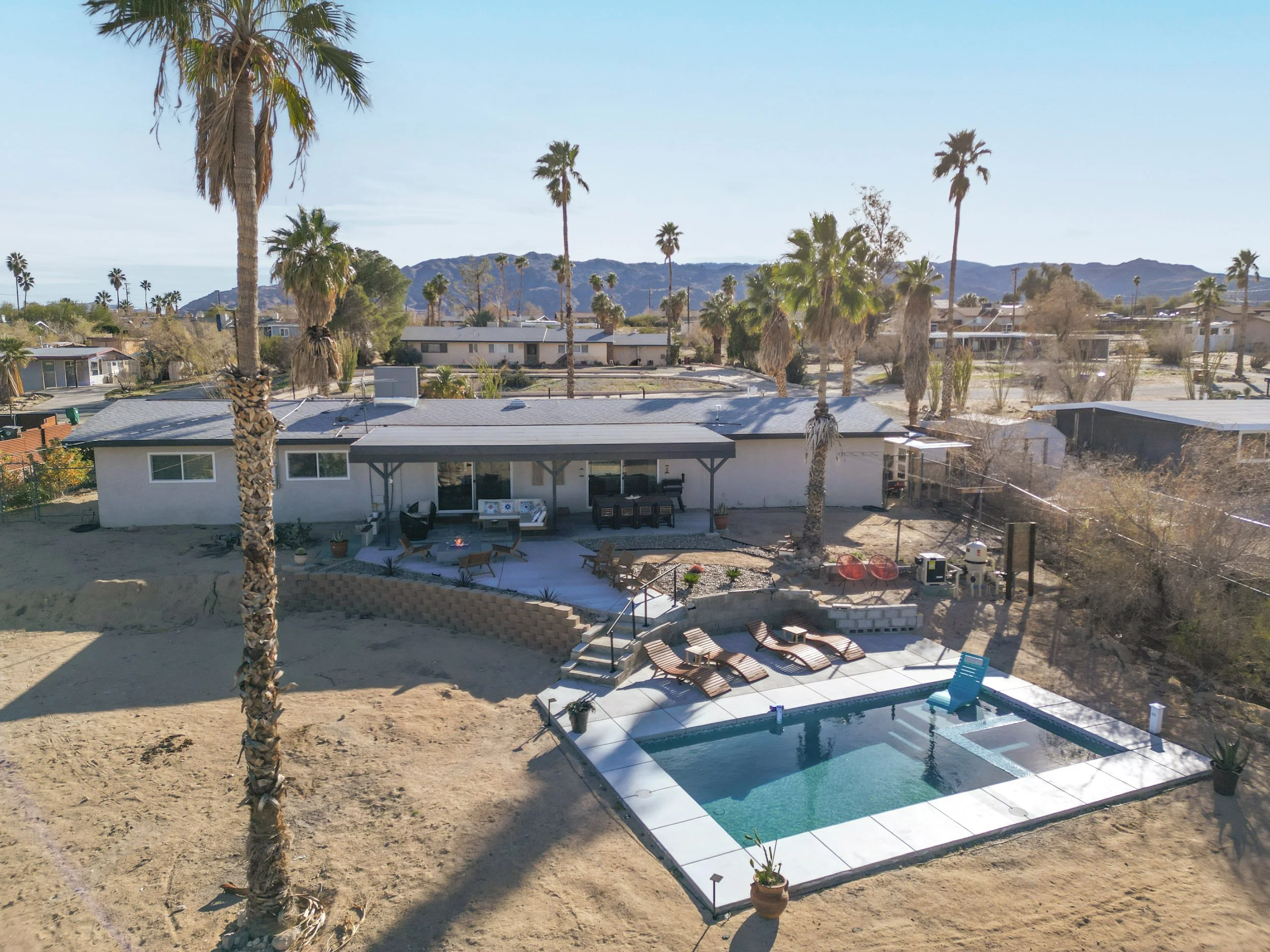 A backyard with a swimming pool, lounge chairs, and palm trees in a desert-like landscape, with houses and mountains in the background.