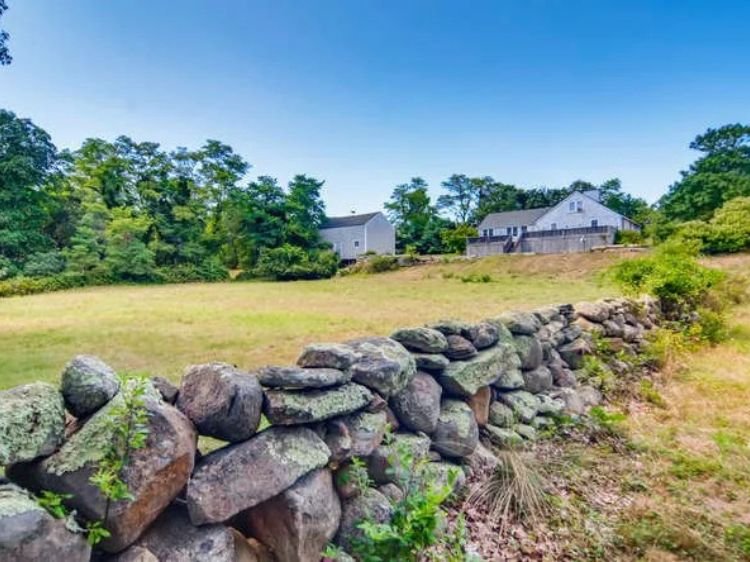 Image of a stonewall with a home in the distance, located in West Tisbury, Martha's Vineyard, North Road. 