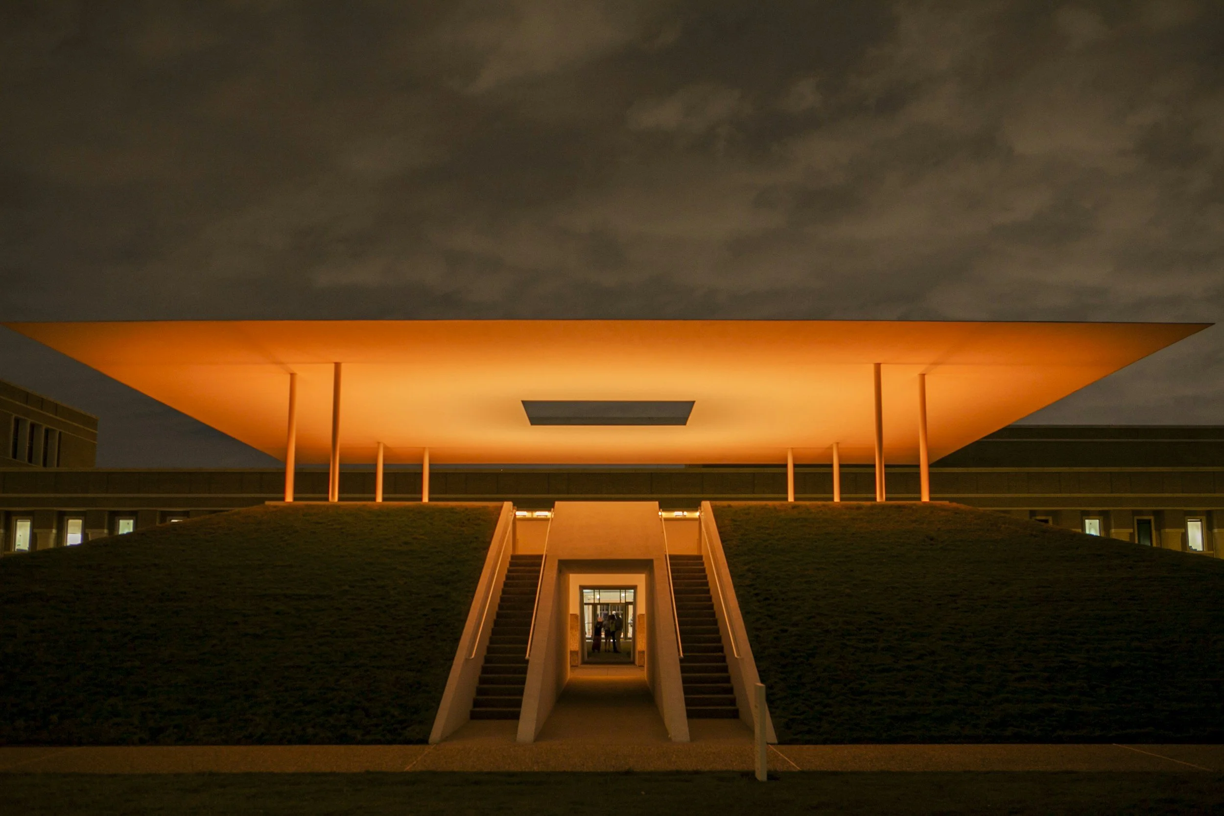 Nighttime view of a modern building with a large flat orange-lit roof supported by thin columns, with stairs leading up to an entrance.