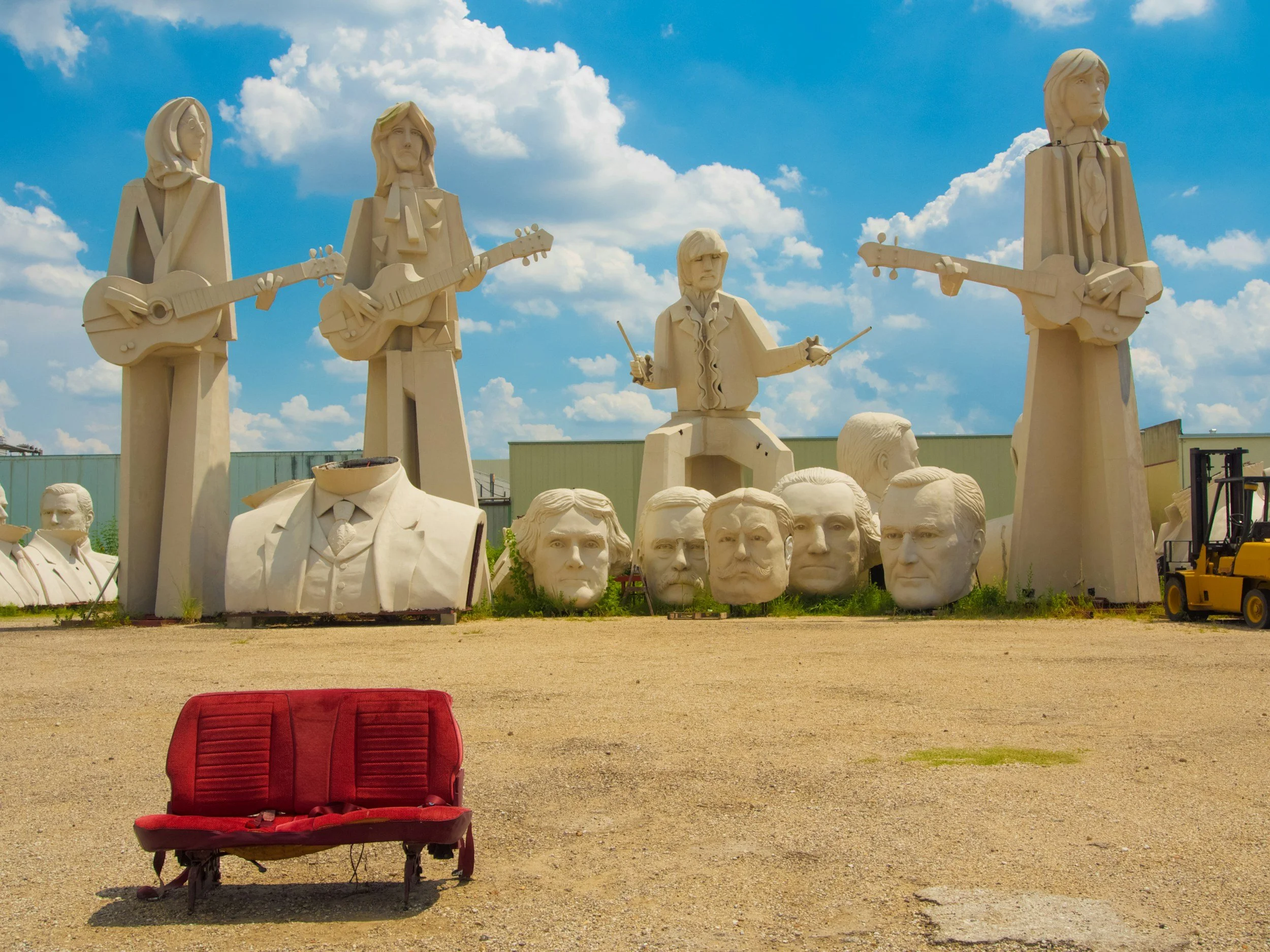 Statues of Mount Rushmore with five large busts, five large statues of musicians playing guitars, a statue of a drummer, and a red couch in the foreground on a sunny day with blue sky and white clouds.