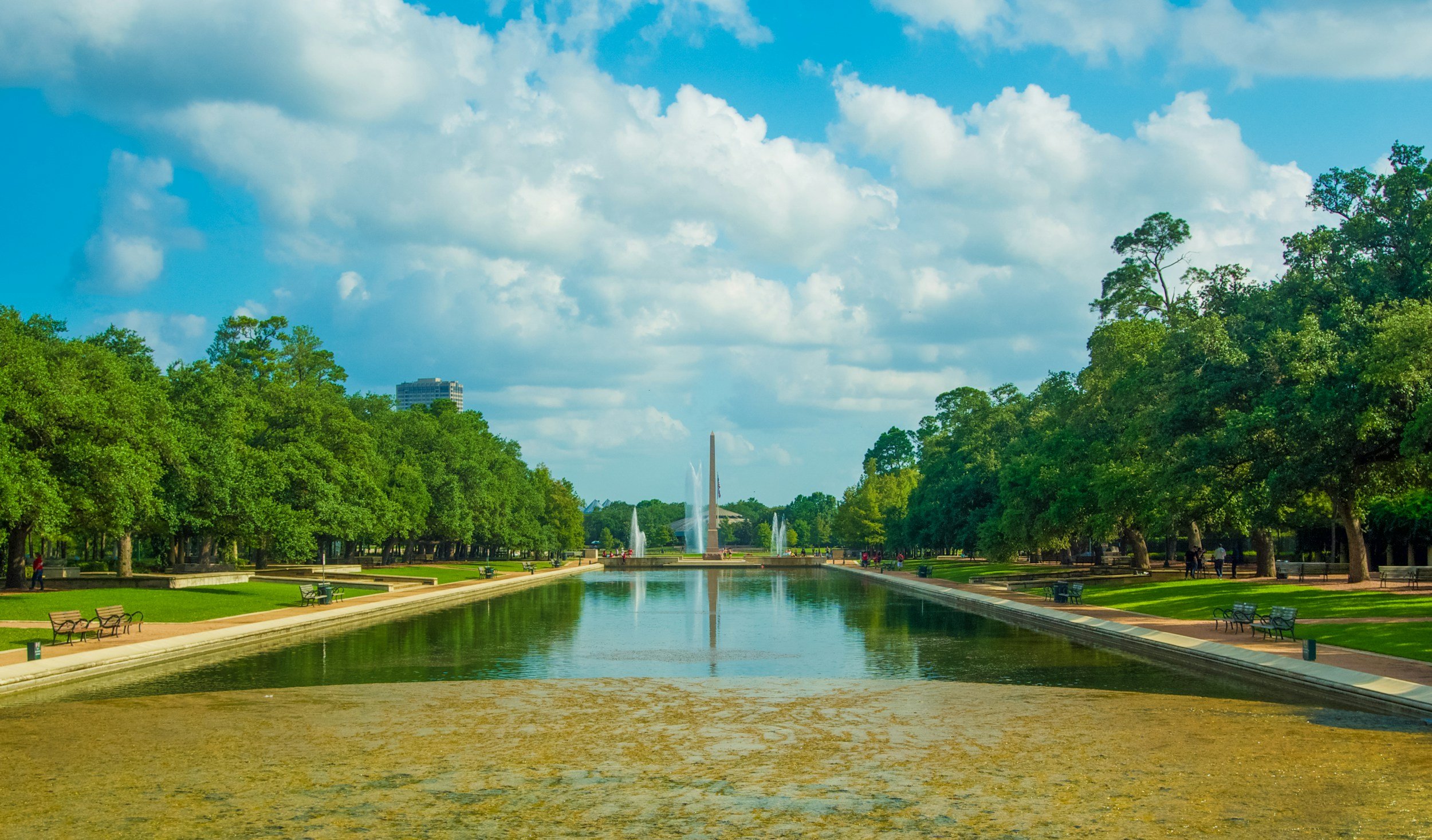 A park with a rectangular reflecting pool, surrounded by green trees. There are benches along the walkway, fountains in the pool, and a tall monument or obelisk in the distance. The sky is partly cloudy with some blue visible.
