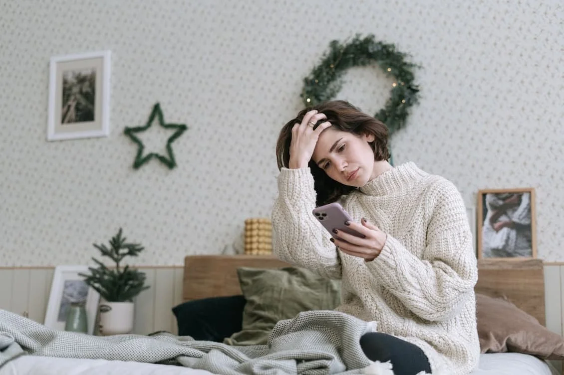 a-woman-in-white-knitted-sweater-sitting-on-bed-while-using-a-smartphone