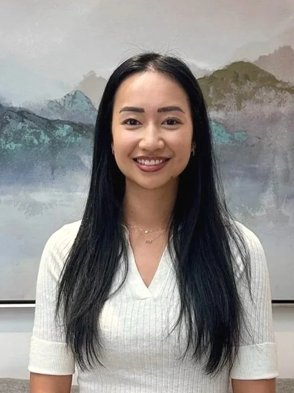 Smiling woman with long black hair wearing a white top in front of a mountain landscape painting