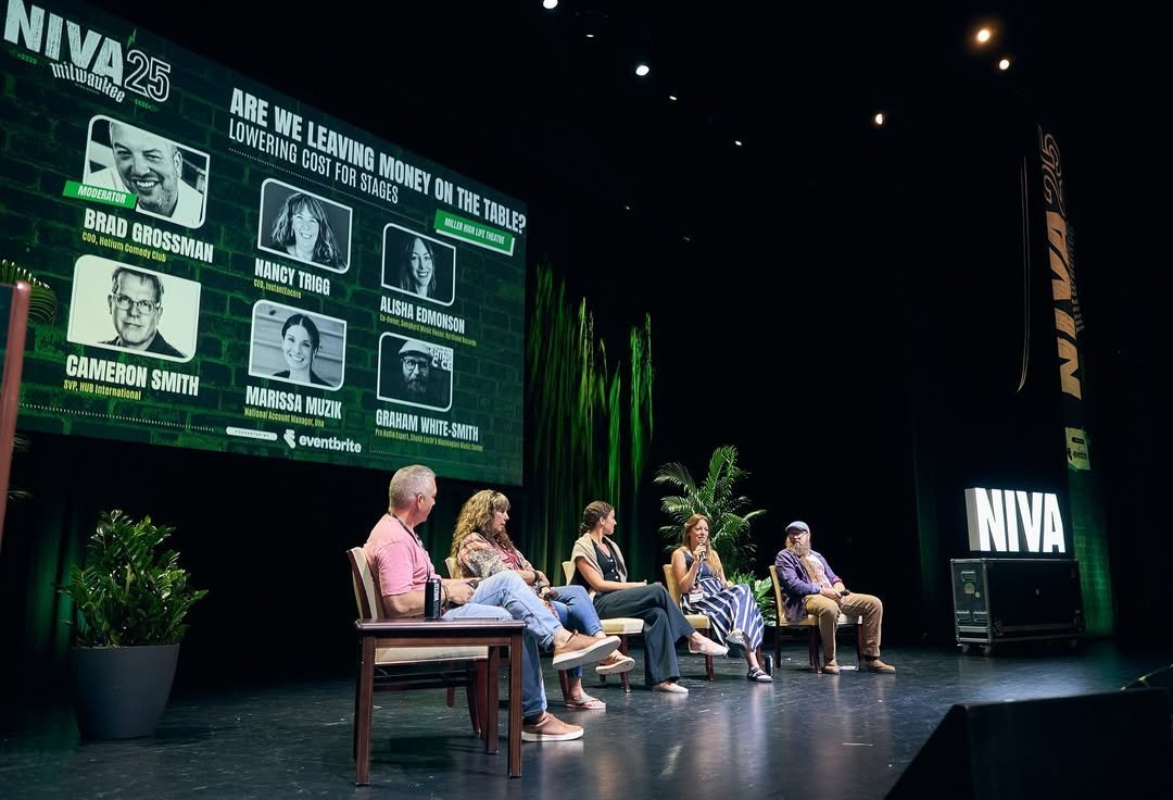 Panel discussion at the NIVA event with five speakers seated on stage, and a large screen behind them displaying names and titles of panelists, along with the NIVA Milwaukee 25th anniversary logo.