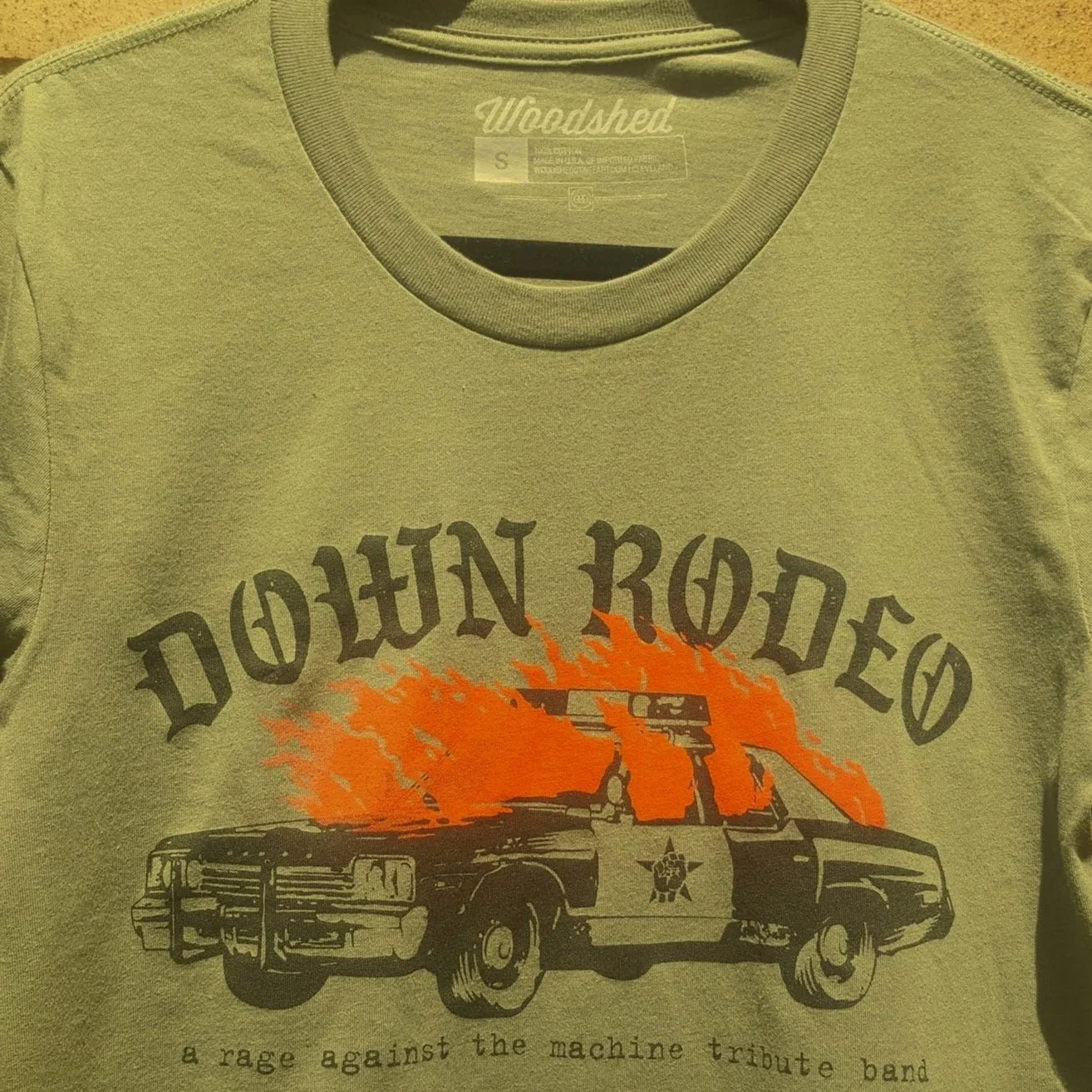 Olive t-shirt with "Down Rodeo" design featuring a burning police car and text "a Rage Against the Machine tribute band."