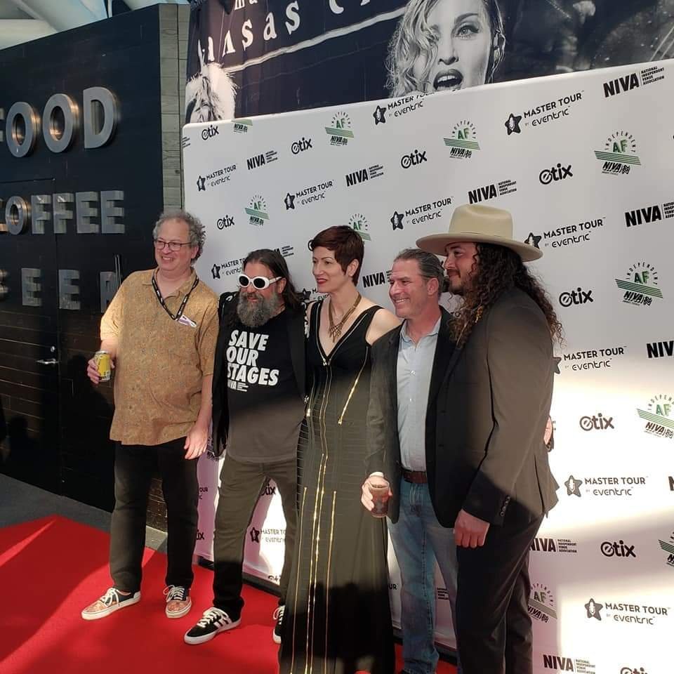 Group of five people posing on a red carpet in front of a backdrop with event logos and names, celebrating a music master tour event.