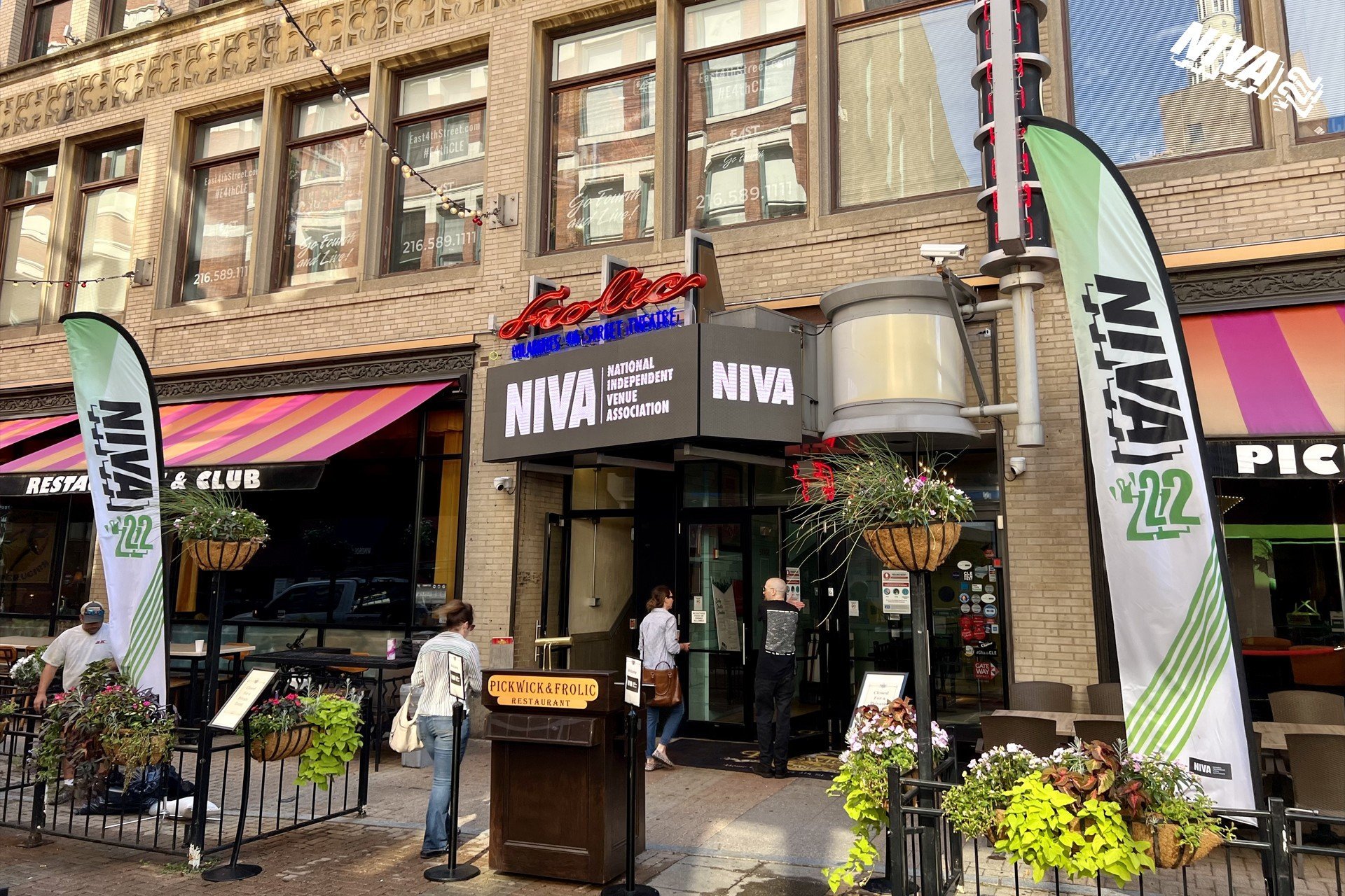 The entrance of a building with signs for the National Independent Venue Association (NIVA) and a restaurant called Pickwick & Frolic, with people walking in and out, decorative plants, and colorful banners outside.