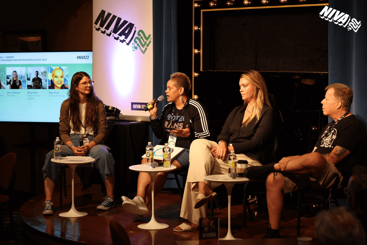 Four women sitting on stage during a panel discussion at a conference, with each woman having a small round table with water bottles in front of her. A woman with short natural hair, black jacket, and striped shirt is speaking into a microphone, whil
