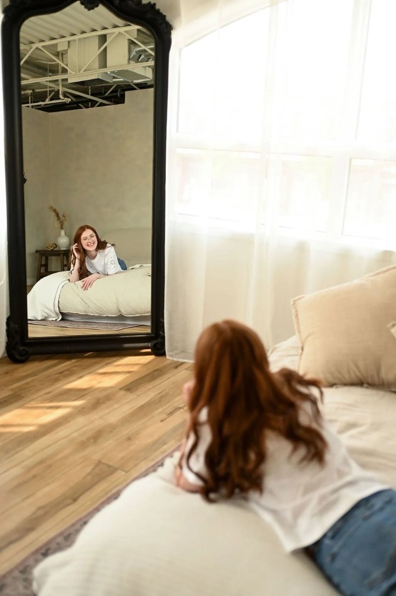 Bright studio scene of woman smiling on bed reflected in large floor mirror.