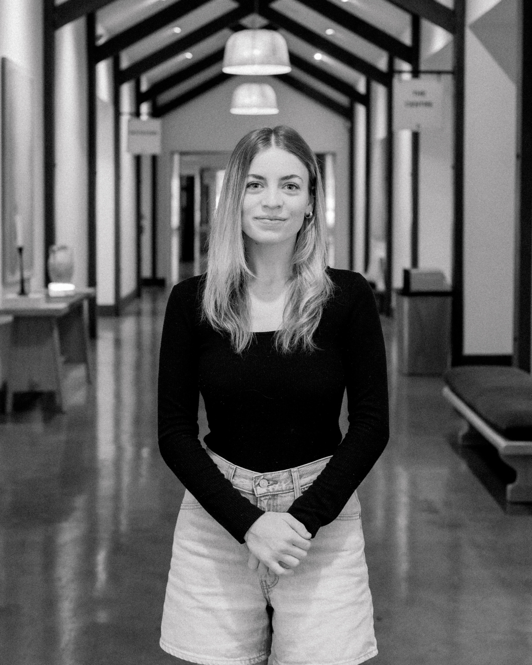 A young woman with blonde wavy hair smiling, wearing a brown jacket, standing indoors.