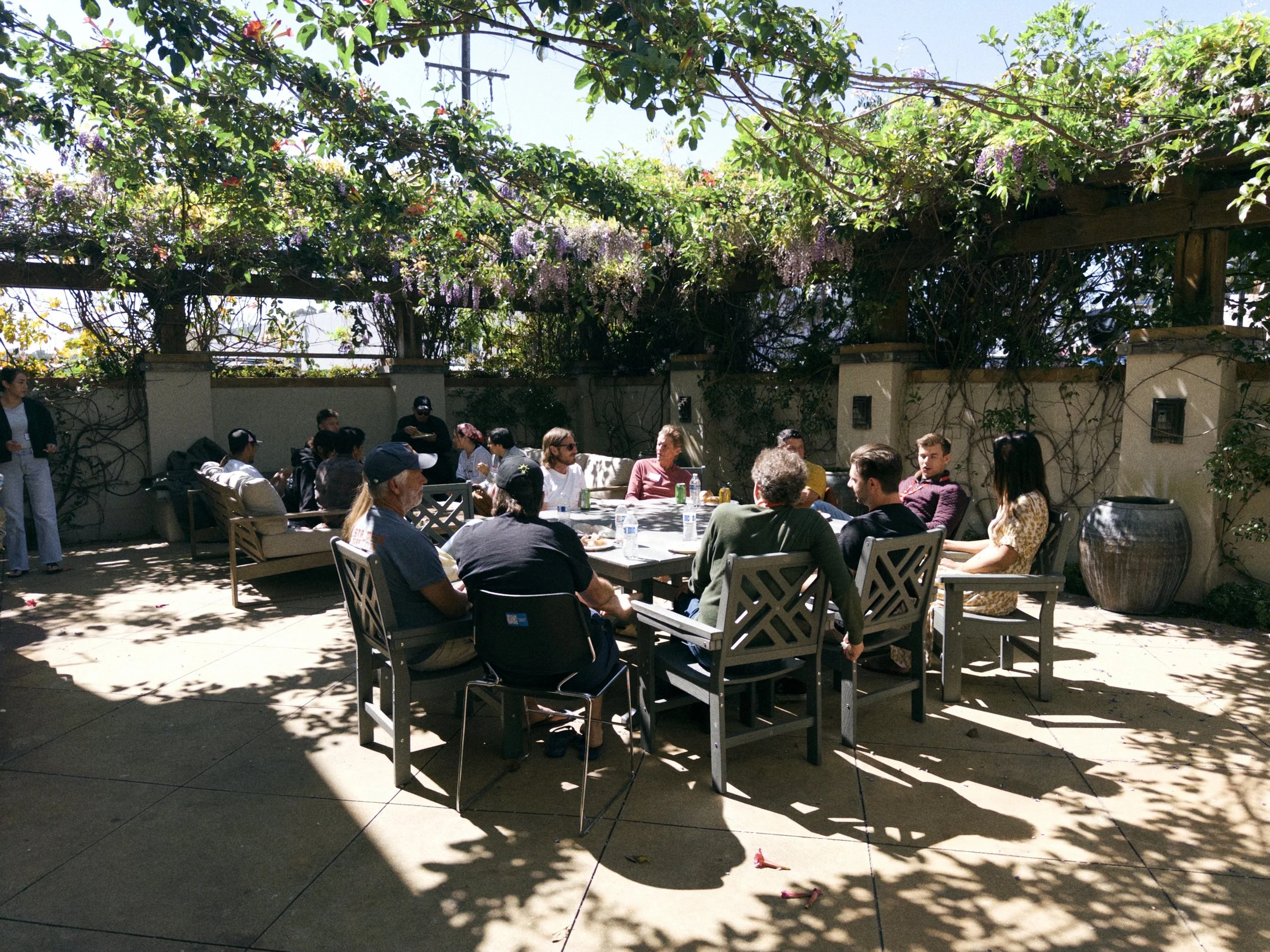 People sitting at tables outdoors in a shaded patio area with lush green plants and flowering vines overhead.