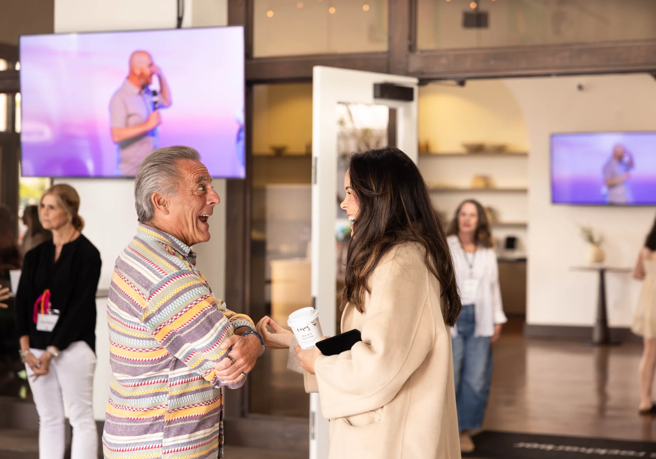 Two people conversing and laughing in a lively, modern indoor space with large screens in the background showing a man speaking on stage. One person is an older man with gray hair wearing a striped shirt, and the other is a younger woman with dark hair, holding a coffee cup and a phone.