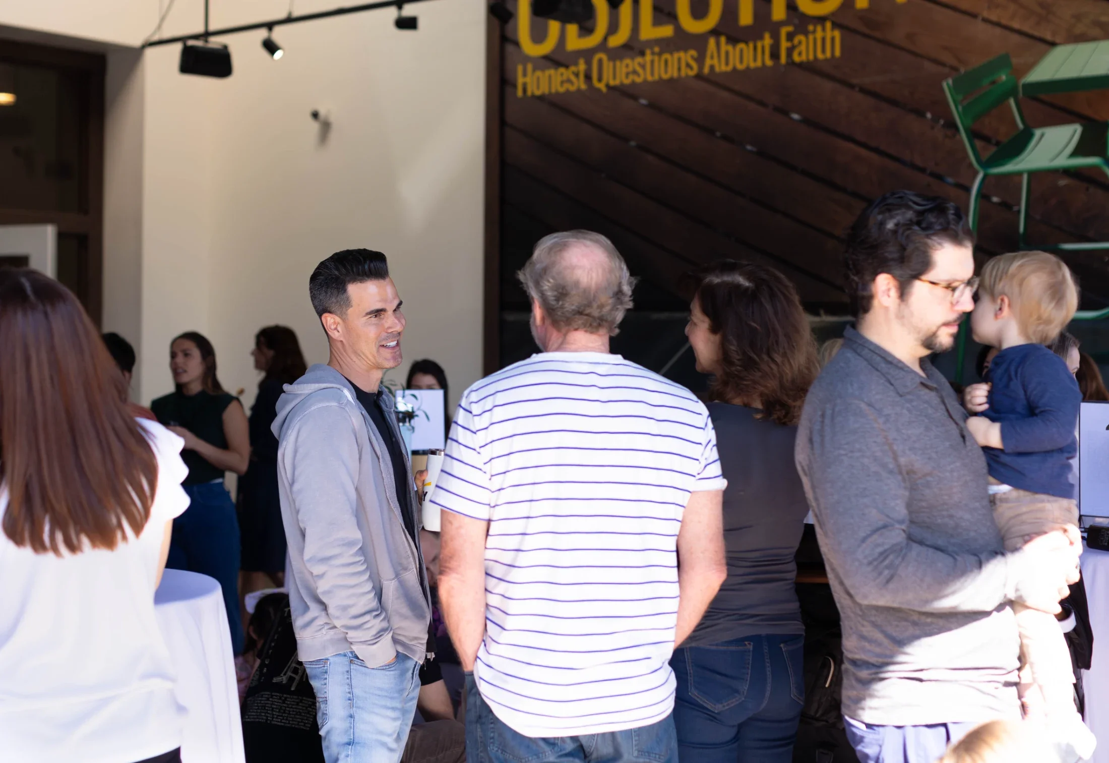 People mingling at an indoor event with a singer in the background, and a large yellow and black sign reading 'ODYSSEY: Honest Questions About Faith' on a wooden wall.