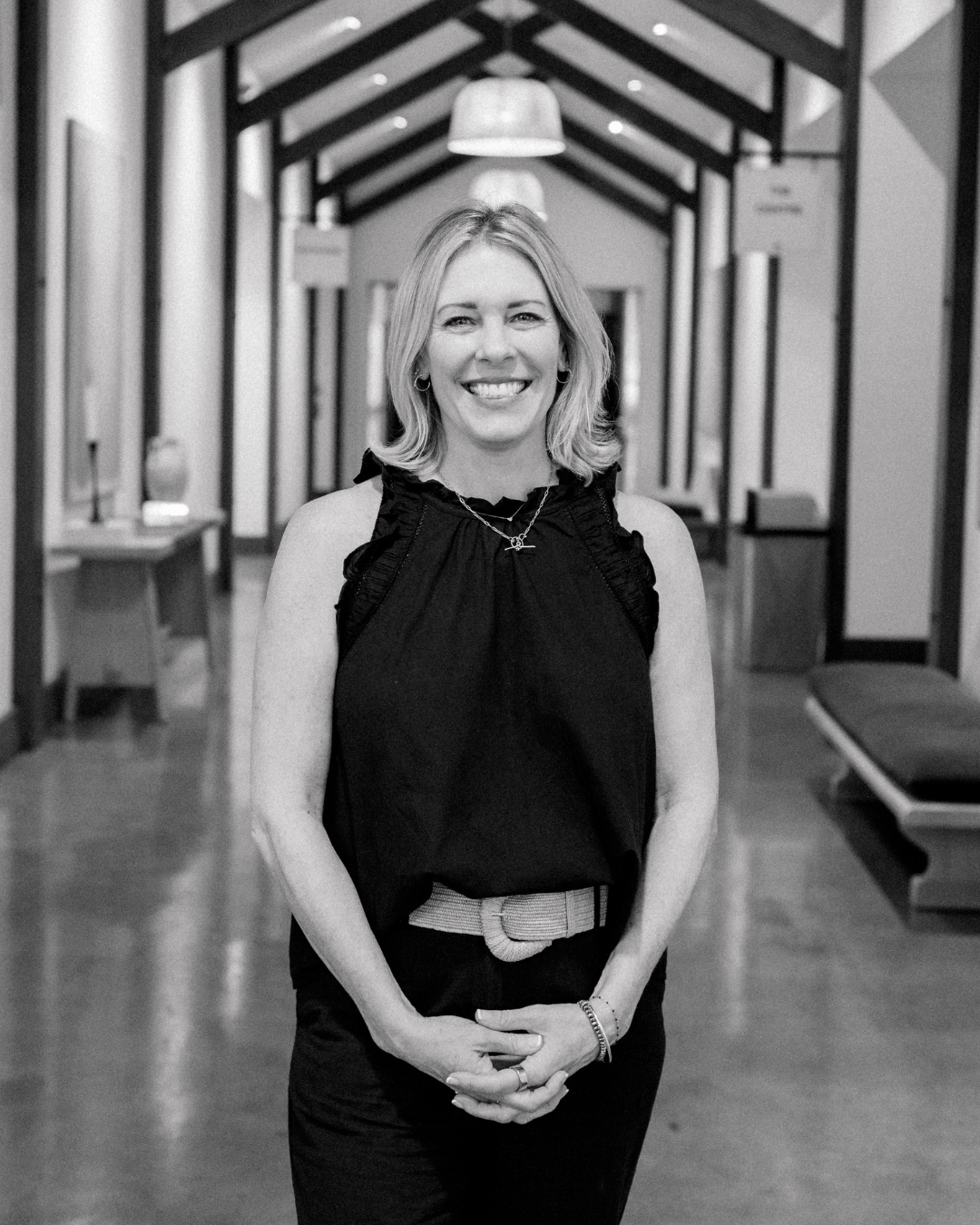 Smiling woman with blonde hair in a black t-shirt standing indoors with windows in the background.