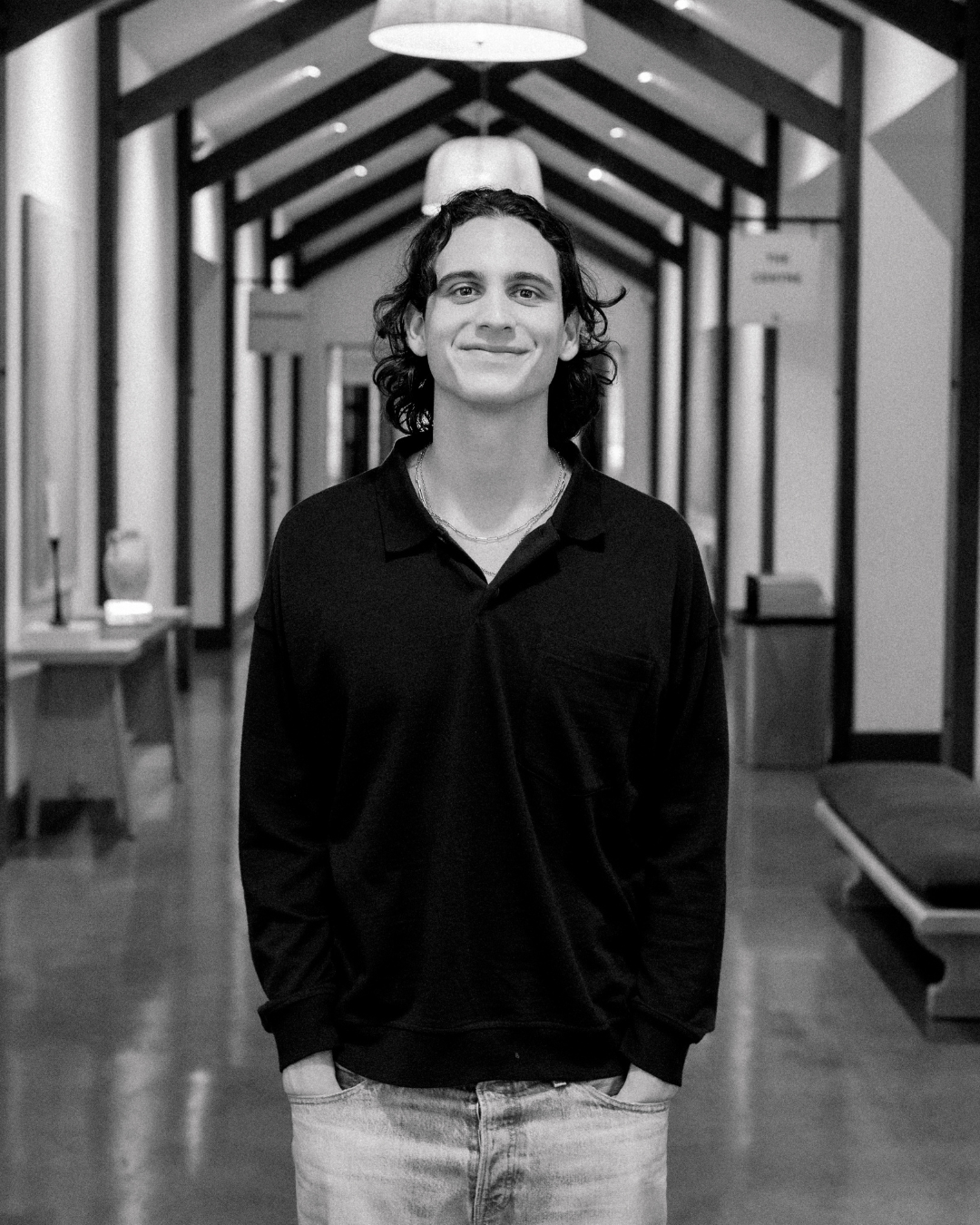 A smiling young man with curly hair wearing a dark jacket, standing indoors with a blurred background.
