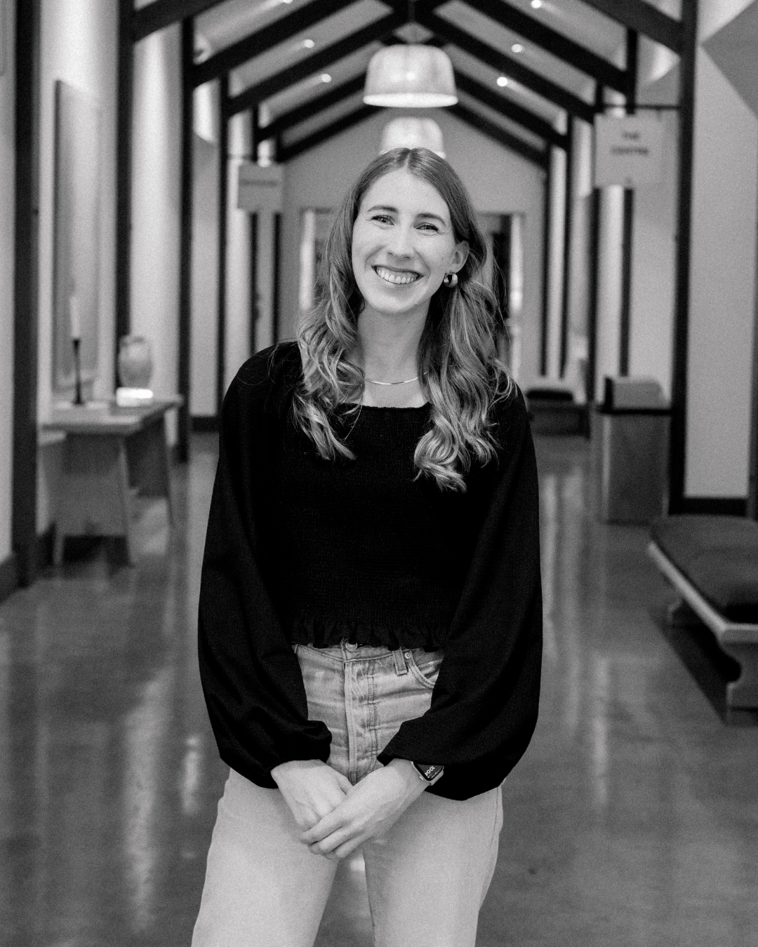 Smiling young woman with long wavy hair wearing a denim jacket and gold earrings, indoors with a dark background and blurred curtains.