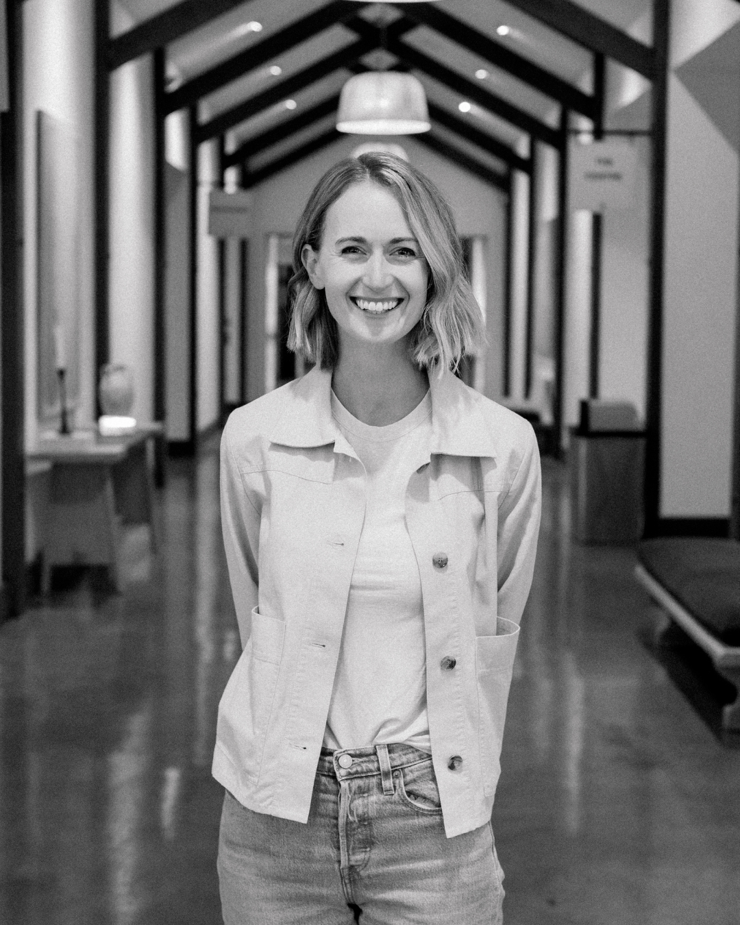 A young woman with shoulder-length brown hair smiles at the camera, wearing a denim jacket over a black top, with a blurred background and sunlight on the right side.