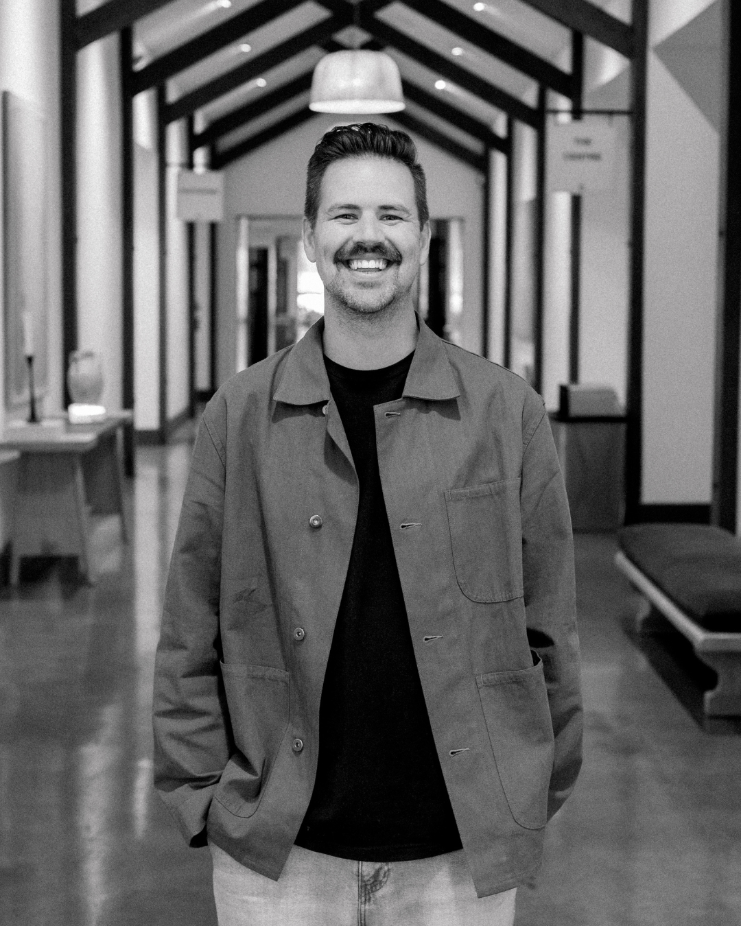 A smiling man with short brown hair and a beard, wearing a light blue denim shirt over a white T-shirt, standing outdoors with a blurred background and warm sunlight on the right side.