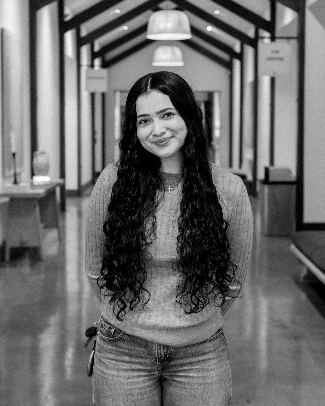 A young woman with long, curly black hair, wearing a black cardigan over a white top, smiling outdoors in front of a building with wooden and glass elements.