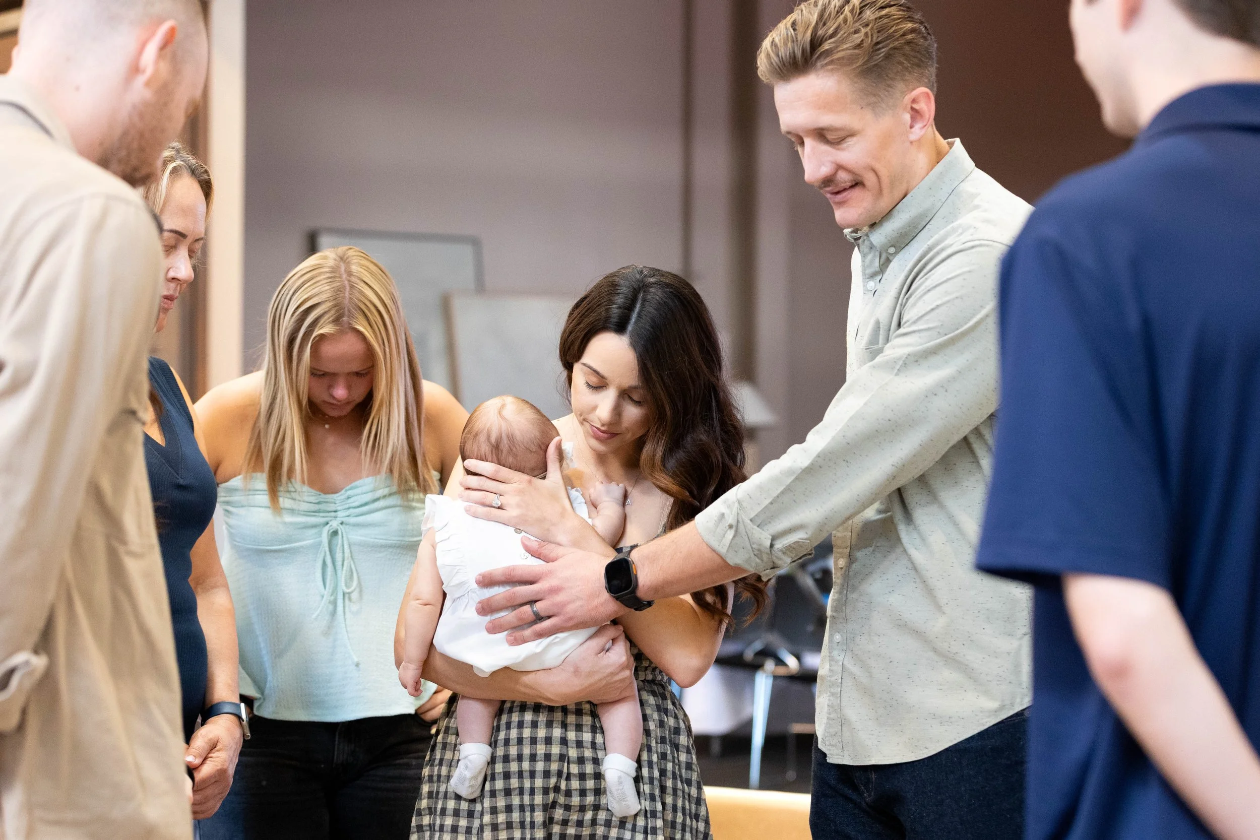 A young child is supported by two adults holding his hands, standing outdoors on grass with trees in the background.
