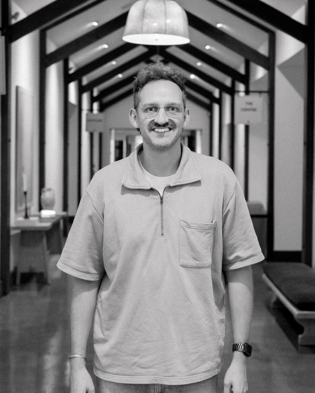 A man with curly hair, glasses, and a beard smiling, wearing a black Carhartt jacket with a white shirt underneath, standing outdoors in warm lighting.