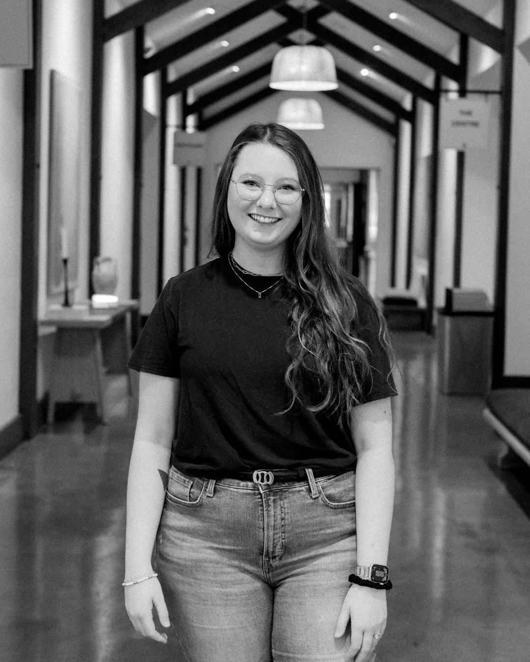 A young woman with long, wavy brown hair and glasses, smiling, wearing a black t-shirt and a light-colored apron with lace detailing, standing indoors with bookshelves in the background.