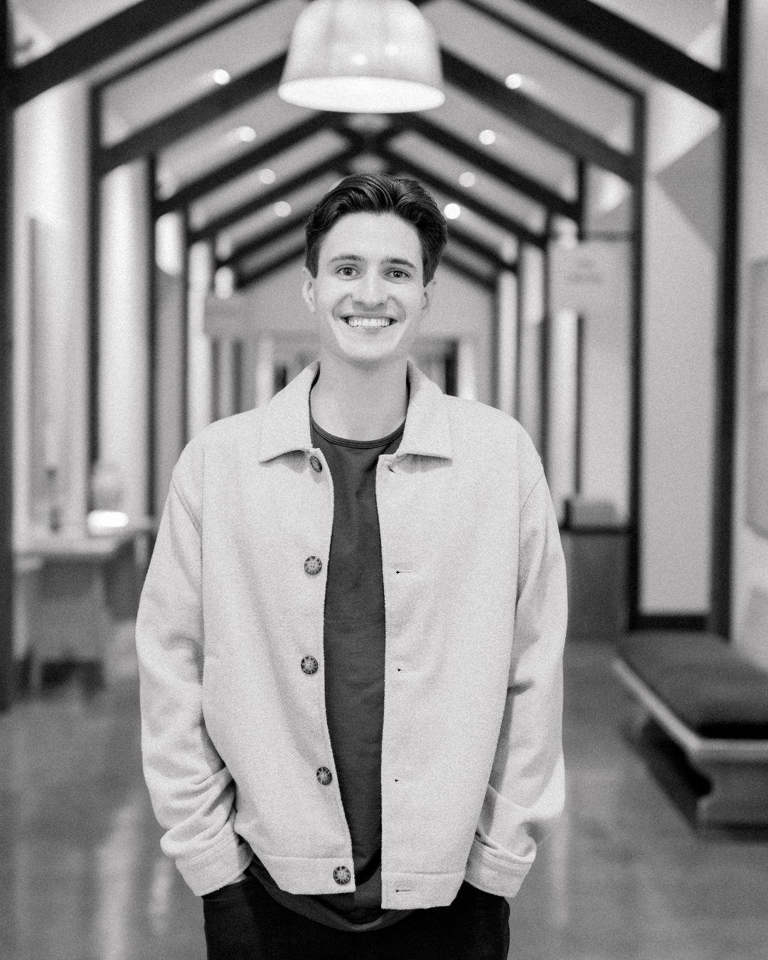 A smiling young man with dark hair wearing a beige coat, standing indoors.