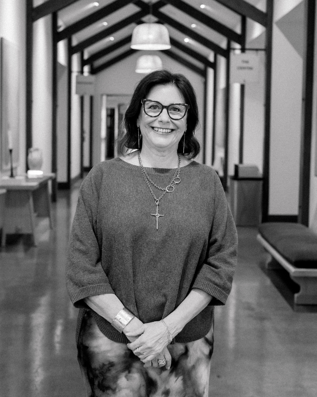 A smiling woman with dark brown hair, wearing glasses, a patterned blouse, a cross necklace, and earrings, standing indoors with a blurred background.