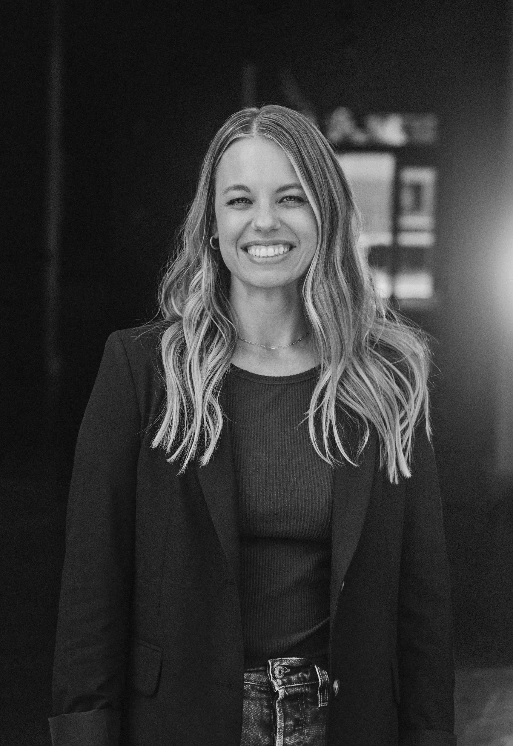 A smiling young woman with blonde hair, wearing a black blazer and a dark green shirt, standing indoors with sunlight in the background.