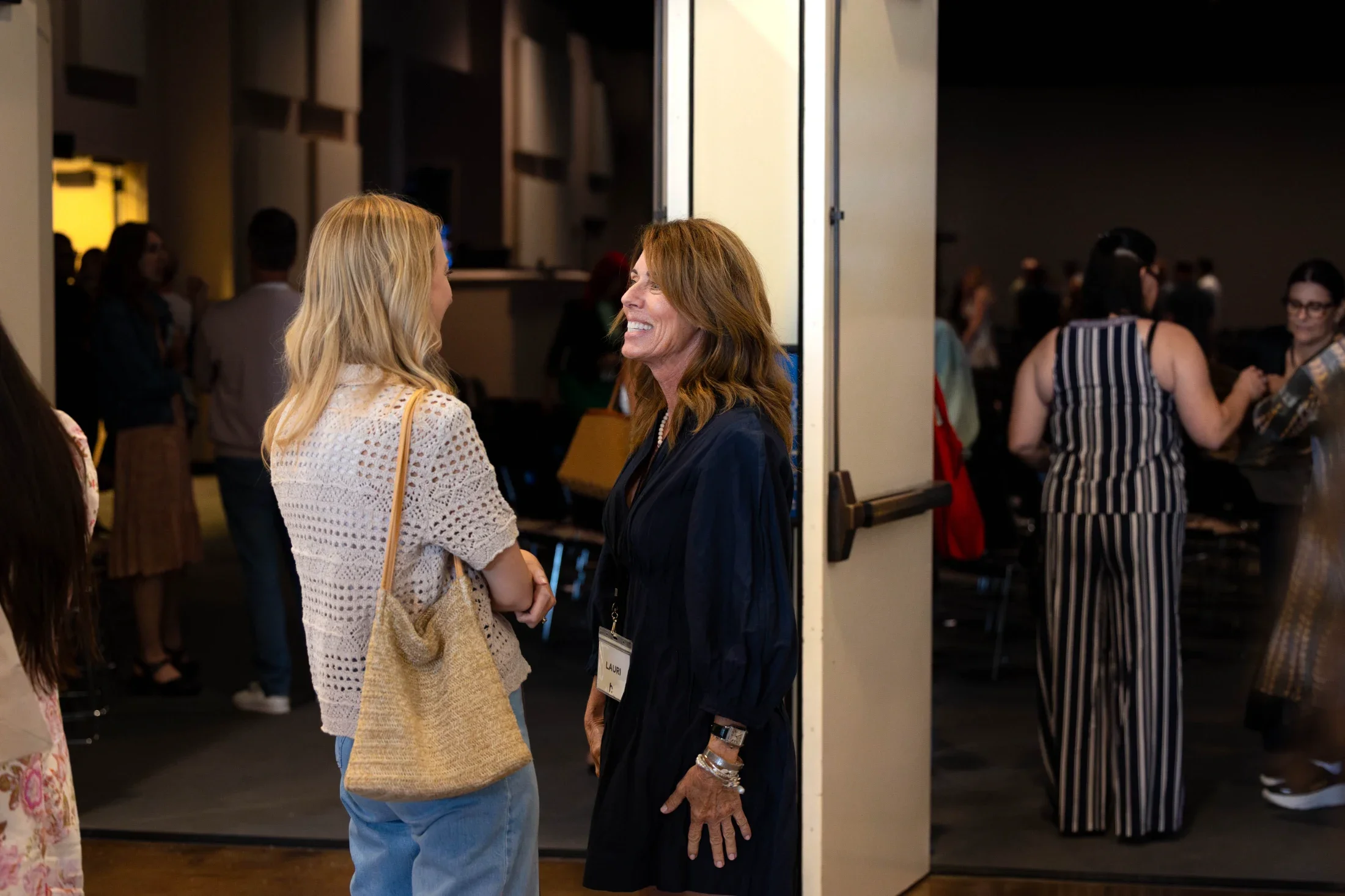 Two women are talking and smiling at each other, standing near a door at a social event or conference. One has blonde hair and is wearing a white crochet top and jeans. The other has reddish hair, wears a dark jacket, and has a name tag that reads "LAUR." In the background, other people are mingling and conversing in a dimly lit room.