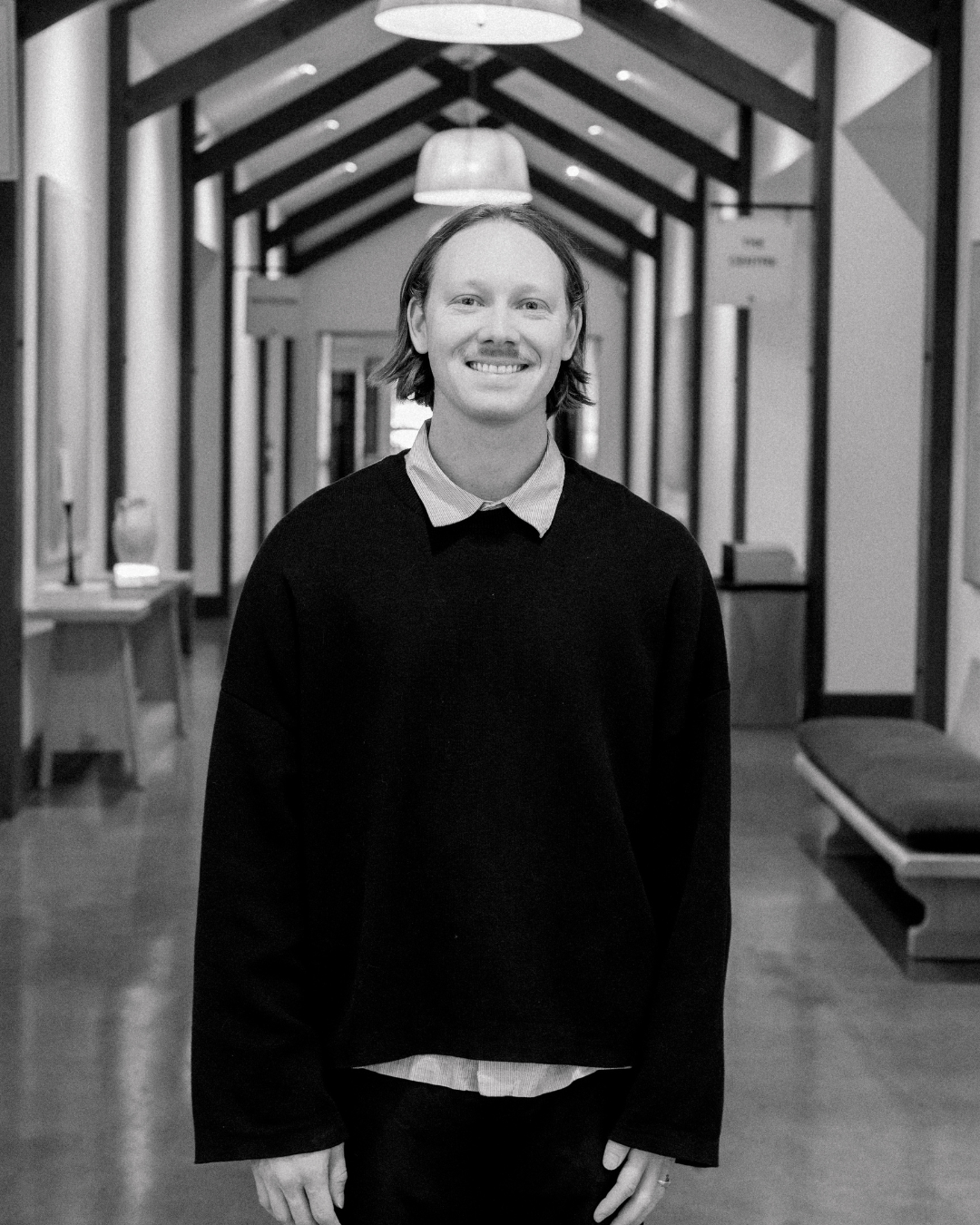 A smiling man with shoulder-length light brown hair, wearing a black short-sleeved button-up shirt, standing outdoors during sunset.