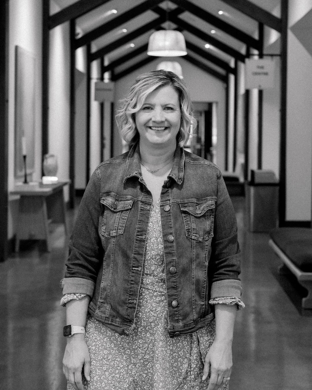 A smiling middle-aged woman with blonde, wavy hair standing indoors in a warmly lit room. She is wearing a navy blue cardigan over a gray camouflage-patterned shirt, a necklace, and a smartwatch on her left wrist.