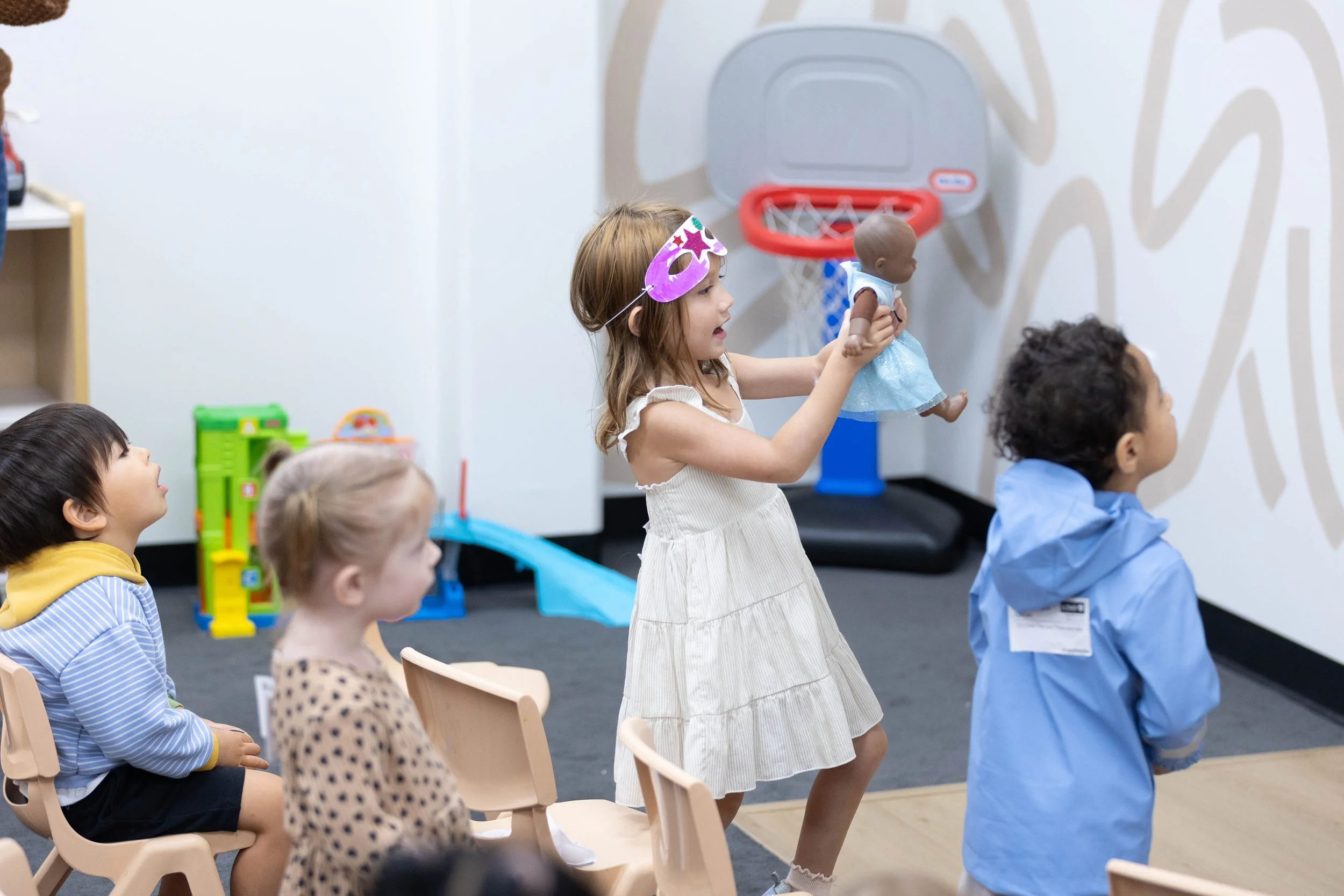 Young girl wearing a light blue dress is drawing with markers at a table with other children, some wearing face masks, in a classroom.